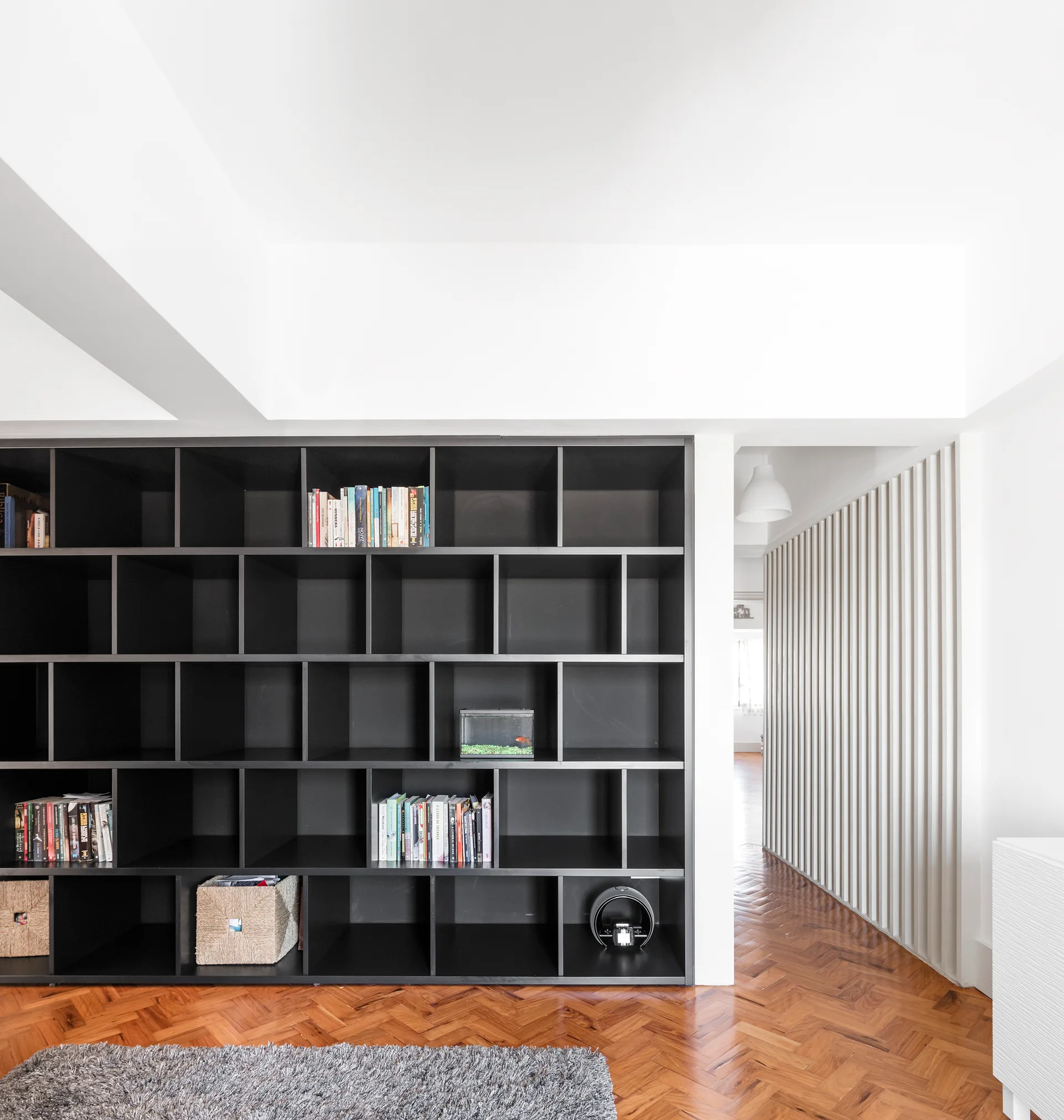 A large black modular bookshelf stands on herringbone patterned wood floors in a bright white room featuring a vertical slatted wall.