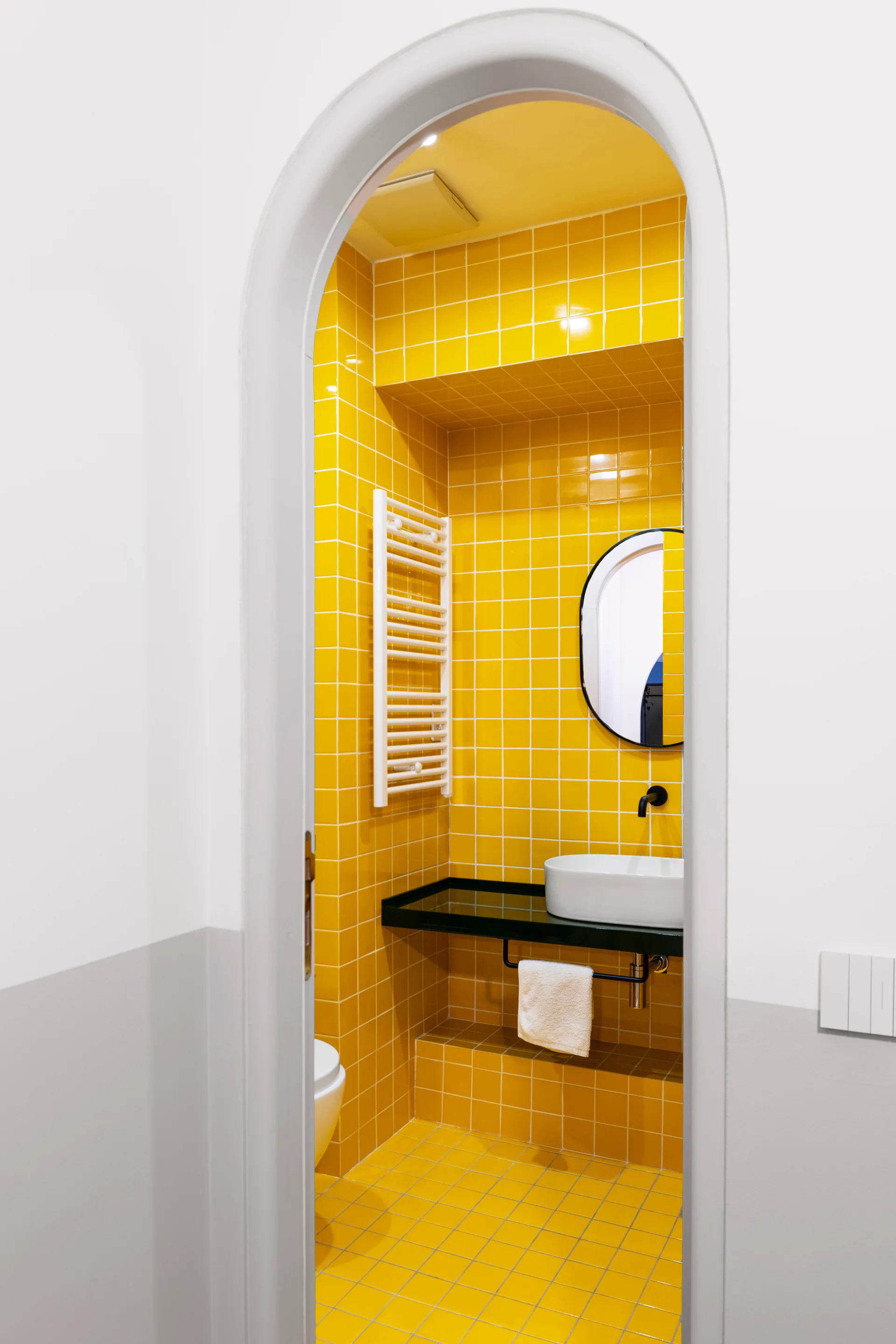 A view through a white arched doorway into a small bathroom with glossy yellow wall tiles, a white vessel sink, and a black faucet.