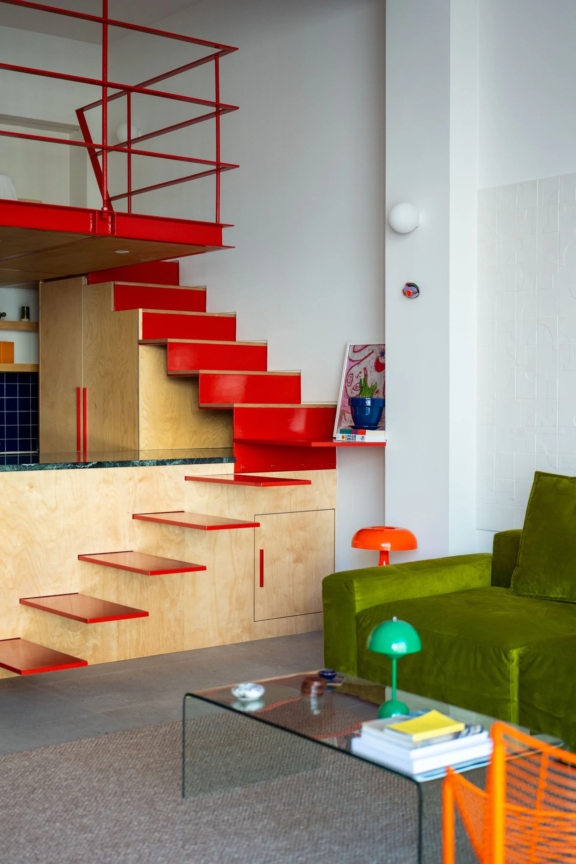 A bright loft apartment featuring a bold red metal staircase with integrated plywood cabinetry, next to a green velvet sofa and glass coffee table.