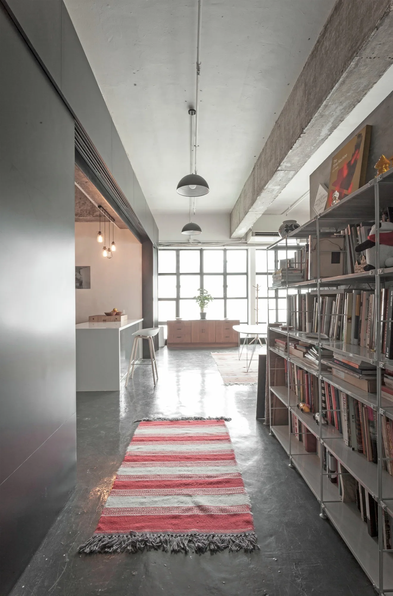 A long industrial loft hallway features a striped runner rug on polished concrete floors, leading past a metal bookcase toward a bright window.