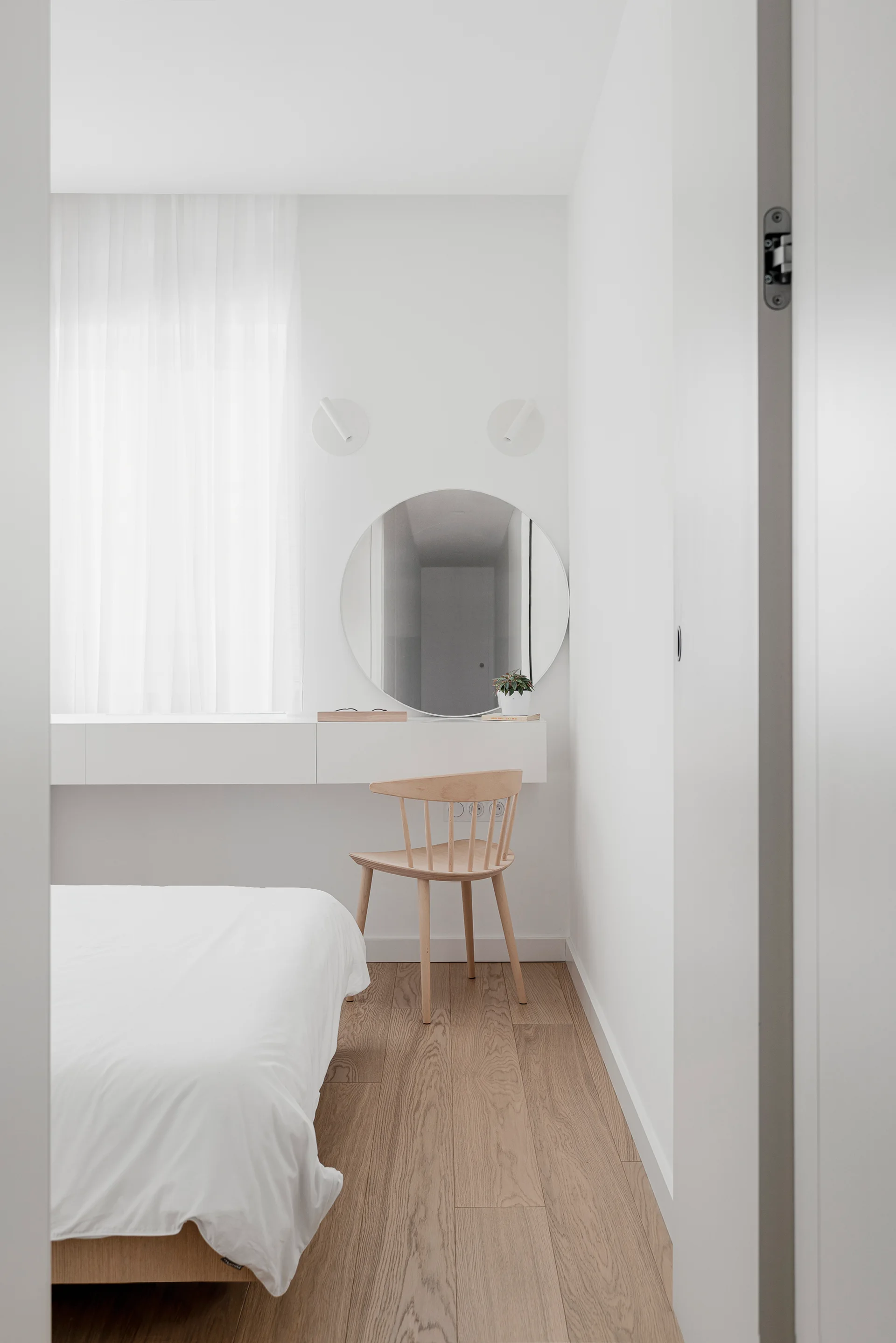 A minimalist bedroom features a wall-mounted white vanity desk, a round mirror, and a light wood spindle chair on hardwood floors.