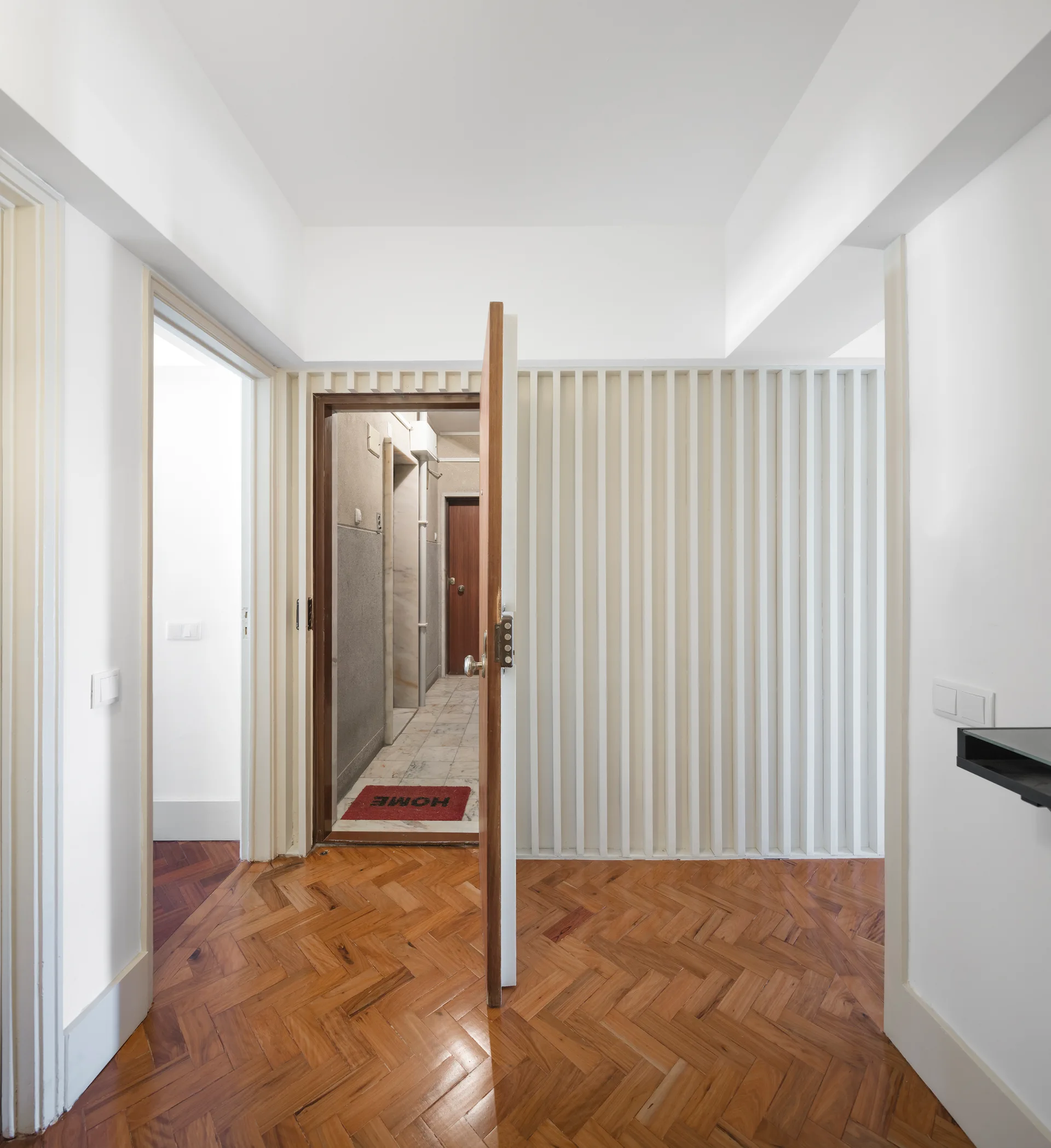 A bright hallway features polished warm wood herringbone floors and a white accent wall textured with vertical slats.