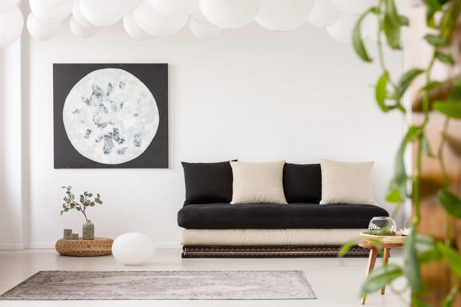 A low-profile black and beige floor sofa sits against a white wall decorated with a large circular moon painting beneath hanging paper lanterns.