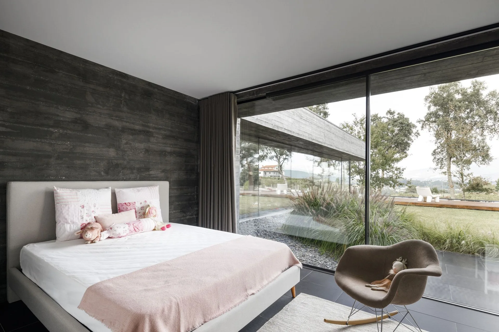 A modern bedroom features an upholstered bed with pink accents, a textured grey concrete wall, and a large glass window looking out onto a green yard.