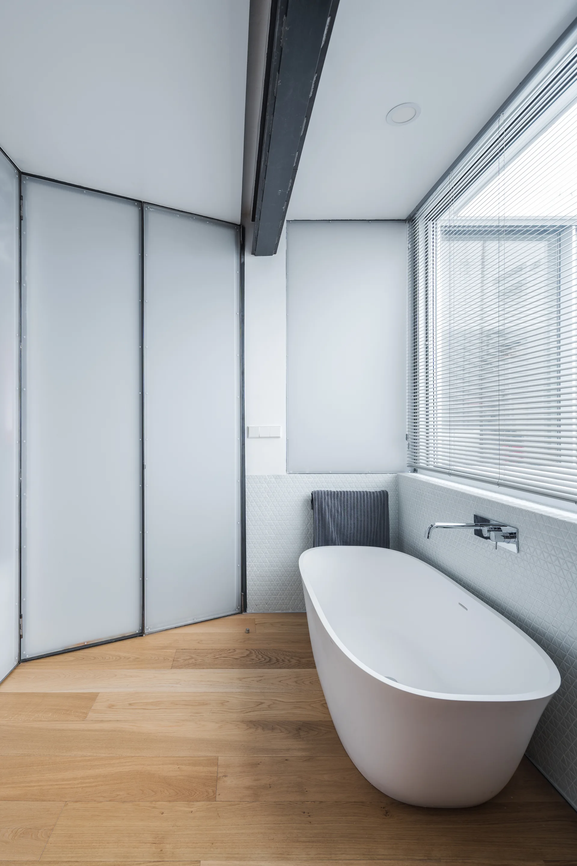 A modern bathroom with a white freestanding bathtub on wood flooring next to a geometric-tiled wall and a large window with blinds.