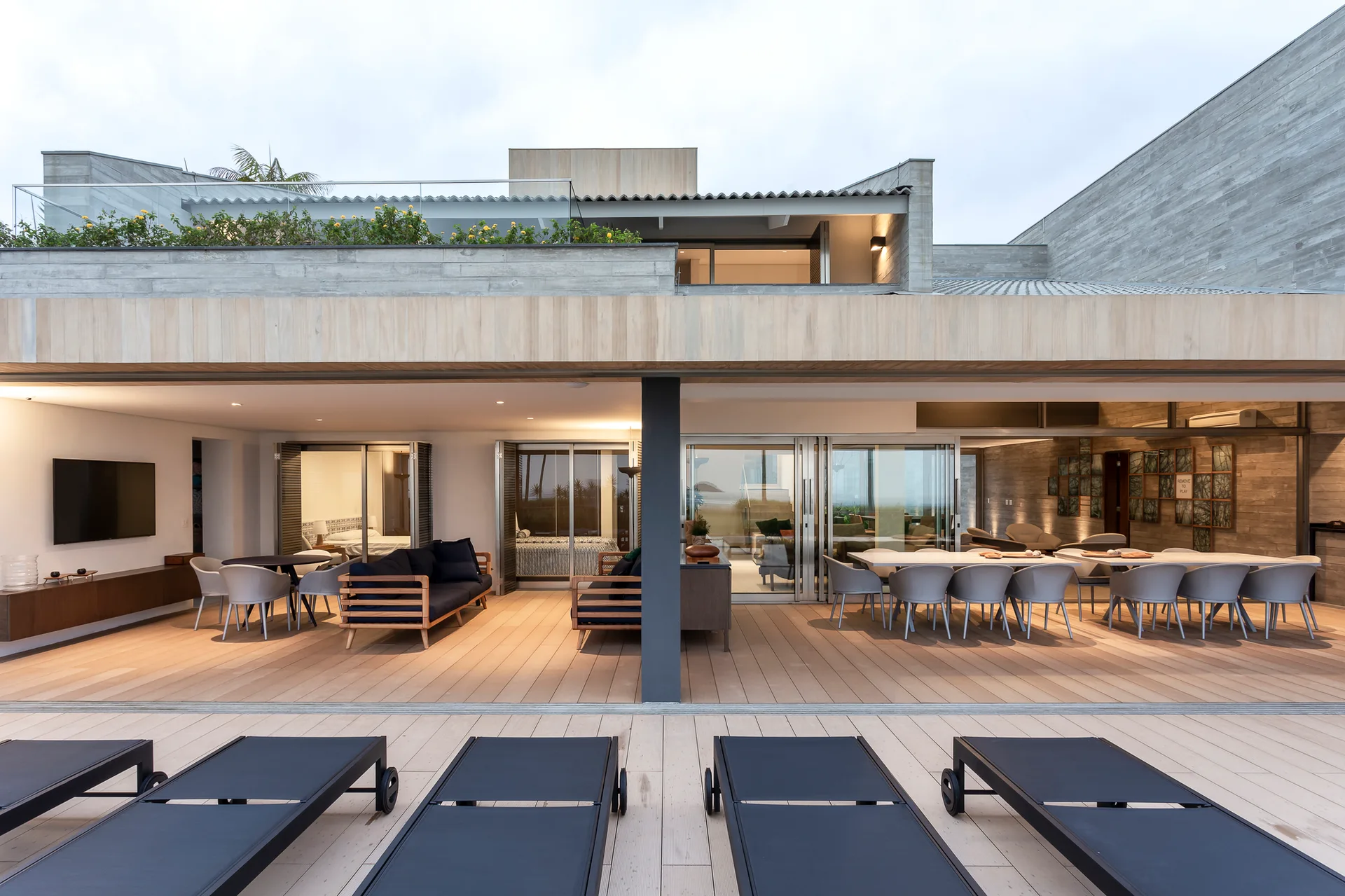 A modern covered patio features light wood decking, outdoor dining tables with grey chairs, low wood-frame sofas, and black sun loungers.