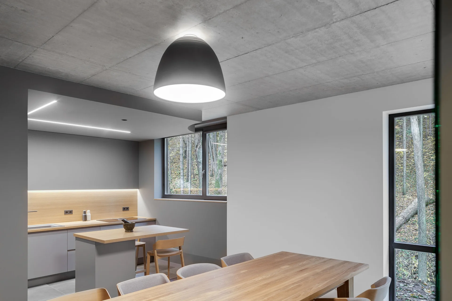 A minimalist kitchen and dining space with grey cabinetry, wood surfaces, an exposed concrete ceiling, and a large window overlooking a forest.