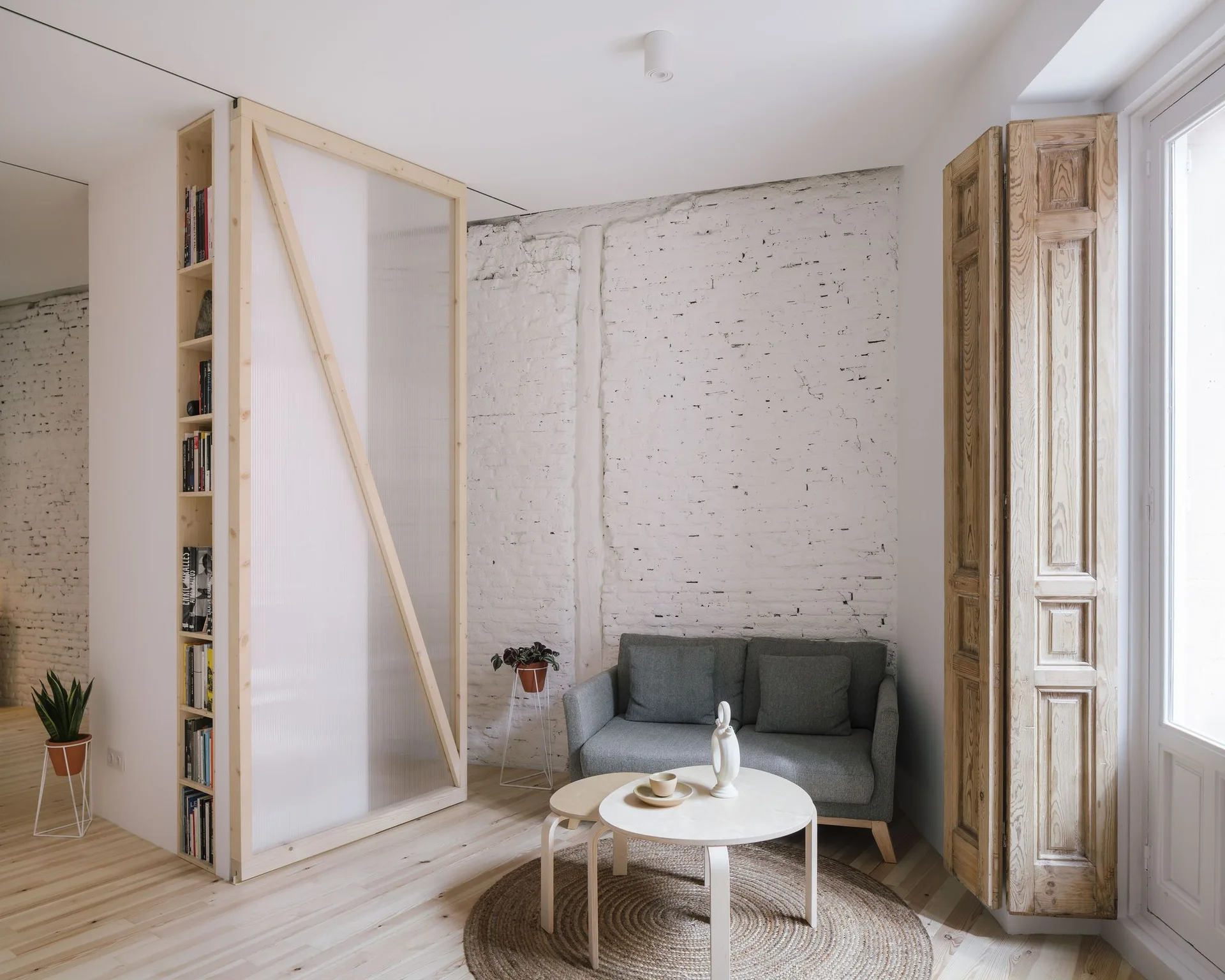 A minimalist living room with a grey sofa, nested wooden coffee tables, and a tall translucent room divider beside a white painted brick wall.