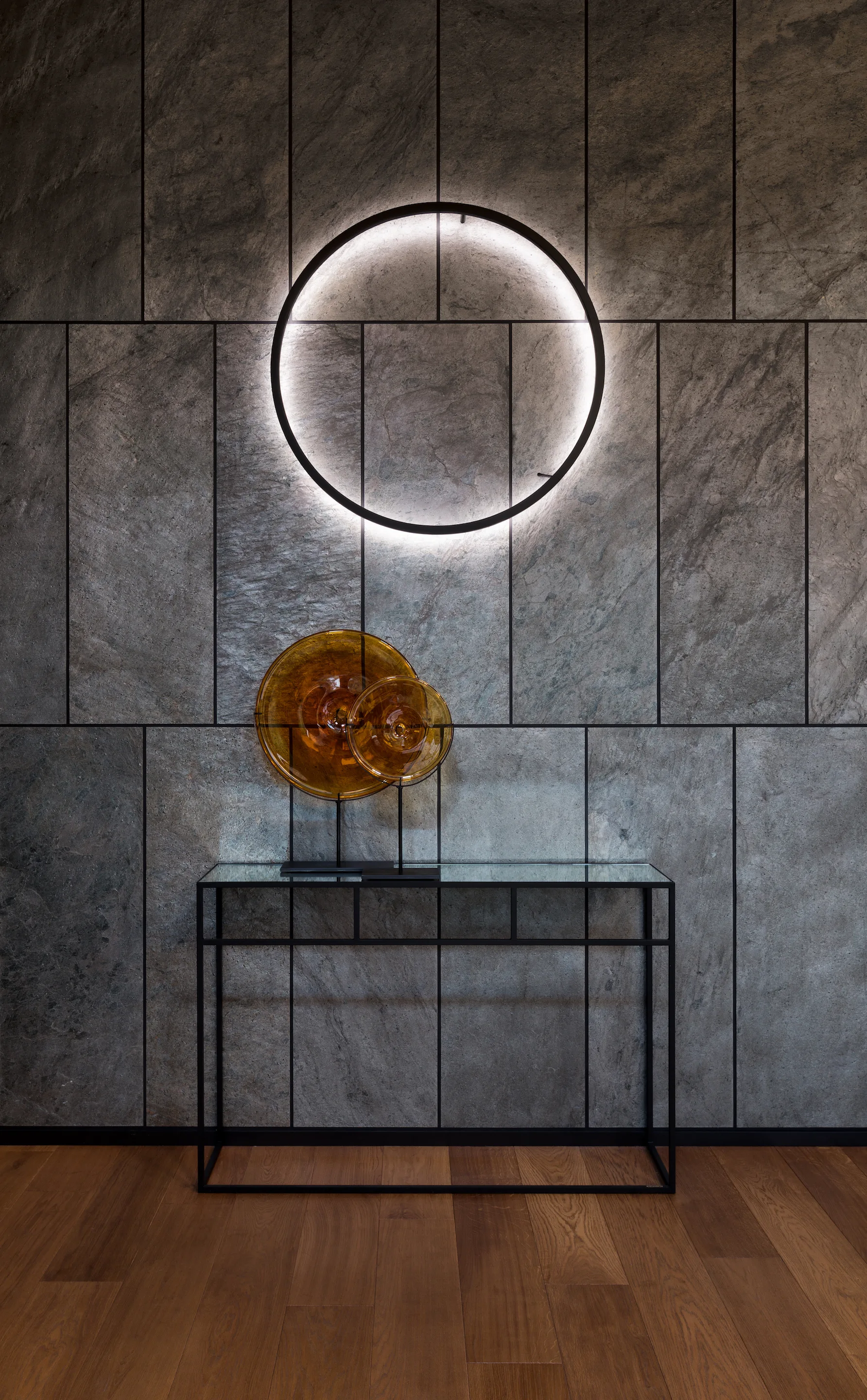 A slim black console table with amber glass sculptures stands against a grey stone tiled wall featuring a glowing circular halo light.