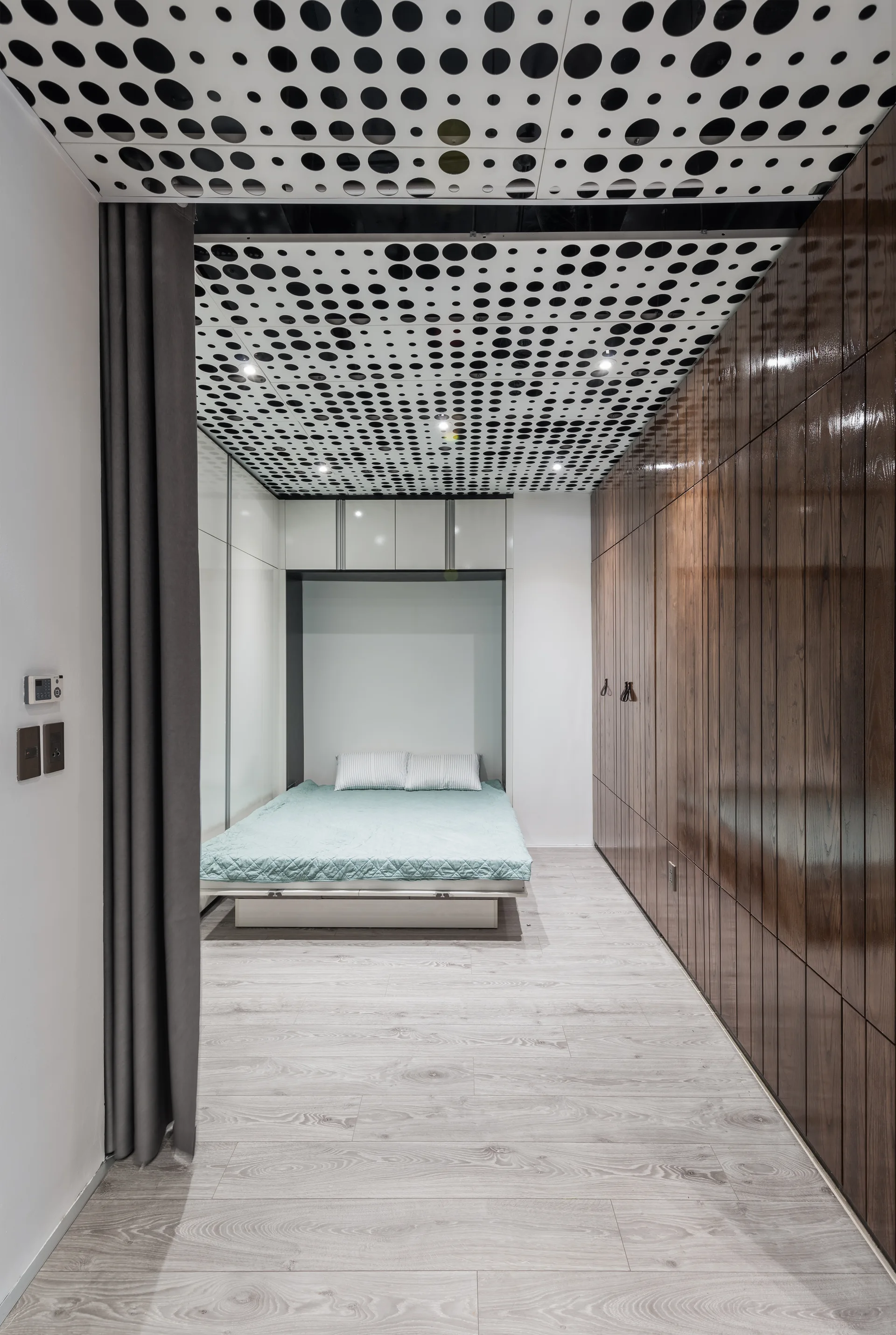 A minimalist bedroom features a low-profile bed with light blue bedding, a glossy dark wood wardrobe wall, and a white ceiling with a black dot pattern.