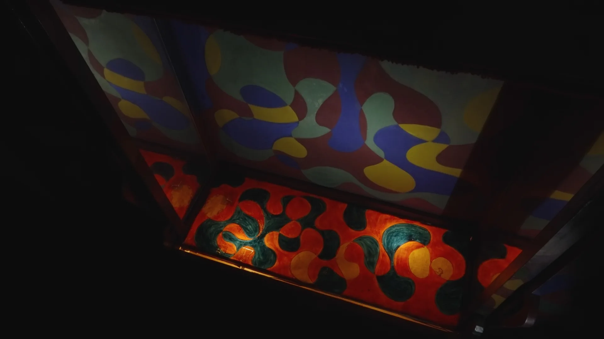 A dimly lit wooden built-in shelf features a colorful hand-painted abstract pattern in red, yellow, blue, and green across its surfaces.