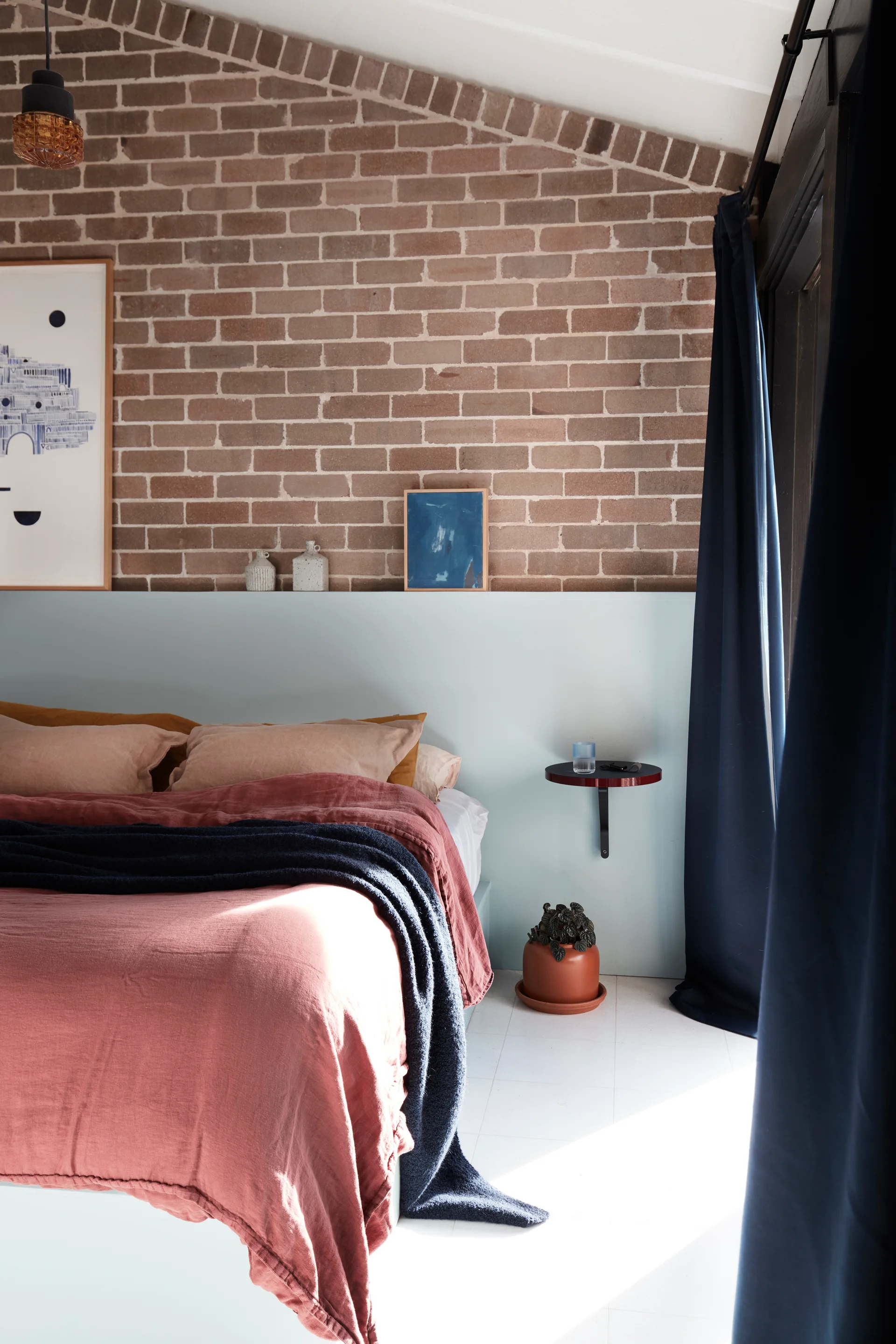 A contemporary bedroom with a rose linen bed set against an exposed brick wall featuring a painted pale blue wainscot ledge and navy curtains.