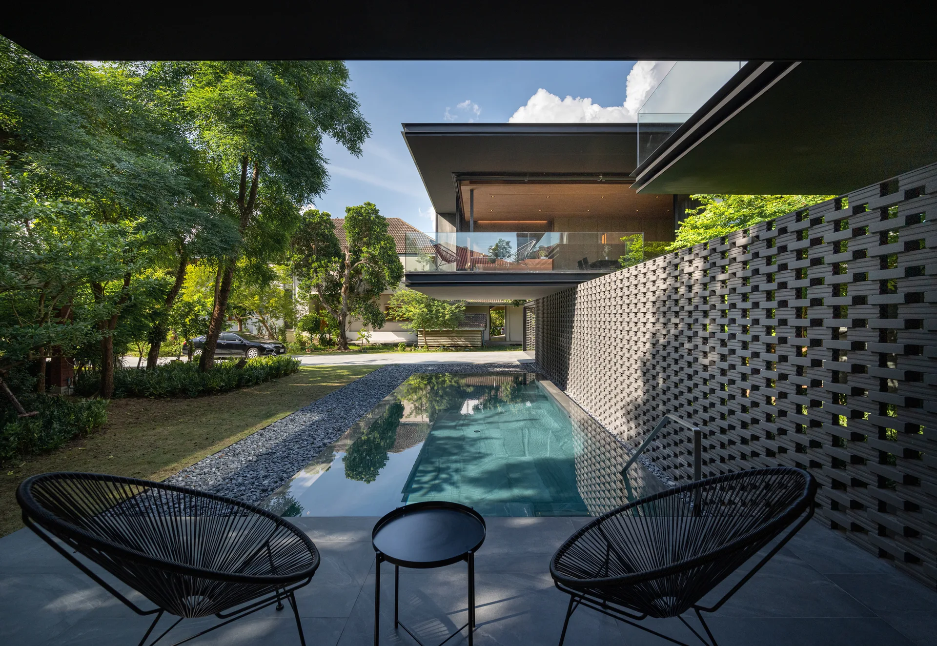 Two black woven chairs on a grey patio overlook a lap pool bordered by a textured brick screen wall and a modern overhanging building.