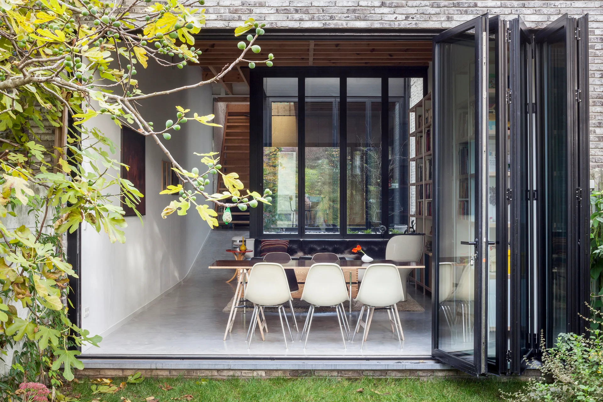 A modern indoor-outdoor dining room with white mid-century chairs opens to a lush green garden through black bifold glass doors.