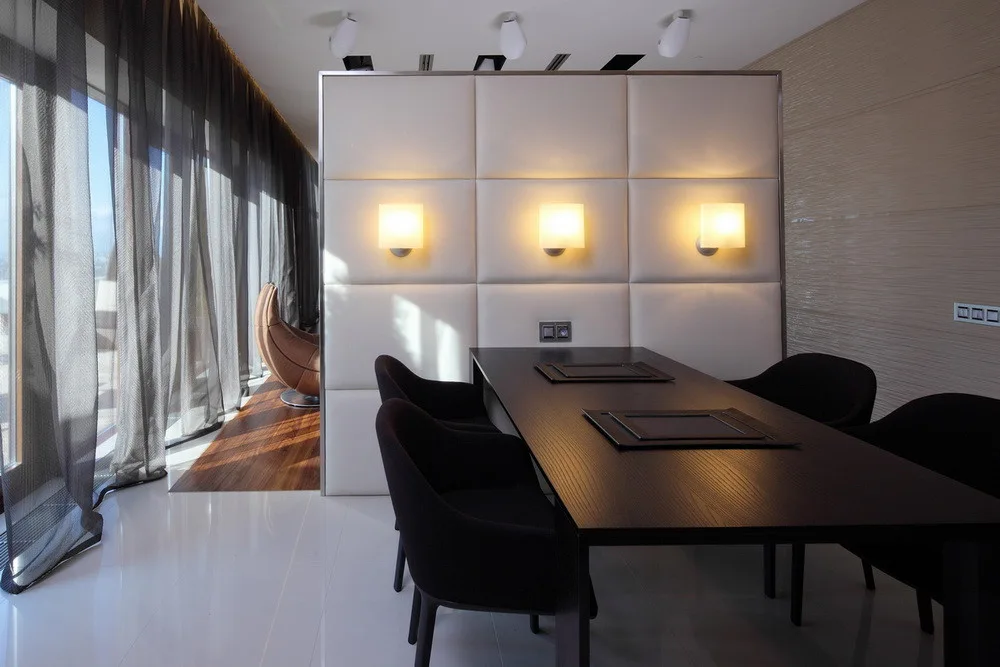 A contemporary dining area featuring a dark wood table with black chairs set against a tall cream upholstered wall panel with three illuminated sconces.