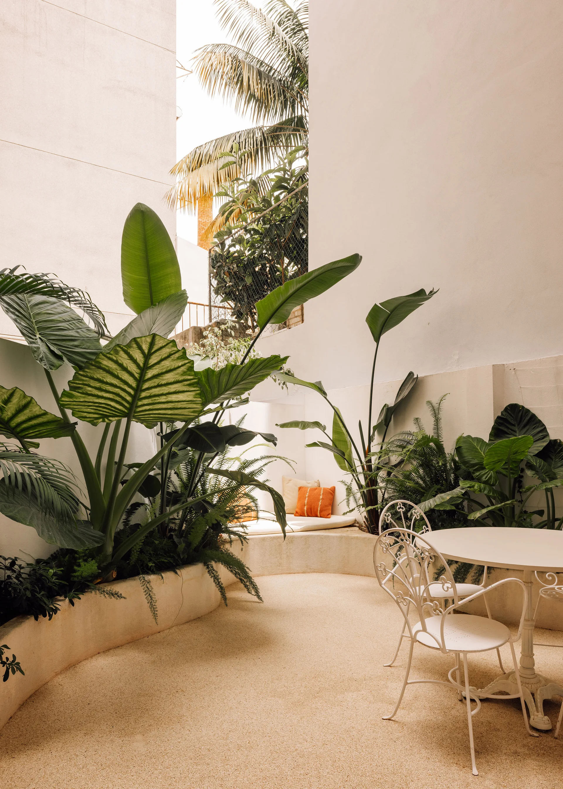 A secluded outdoor courtyard features a white metal bistro set and a curved built-in concrete bench surrounded by lush tropical plants.