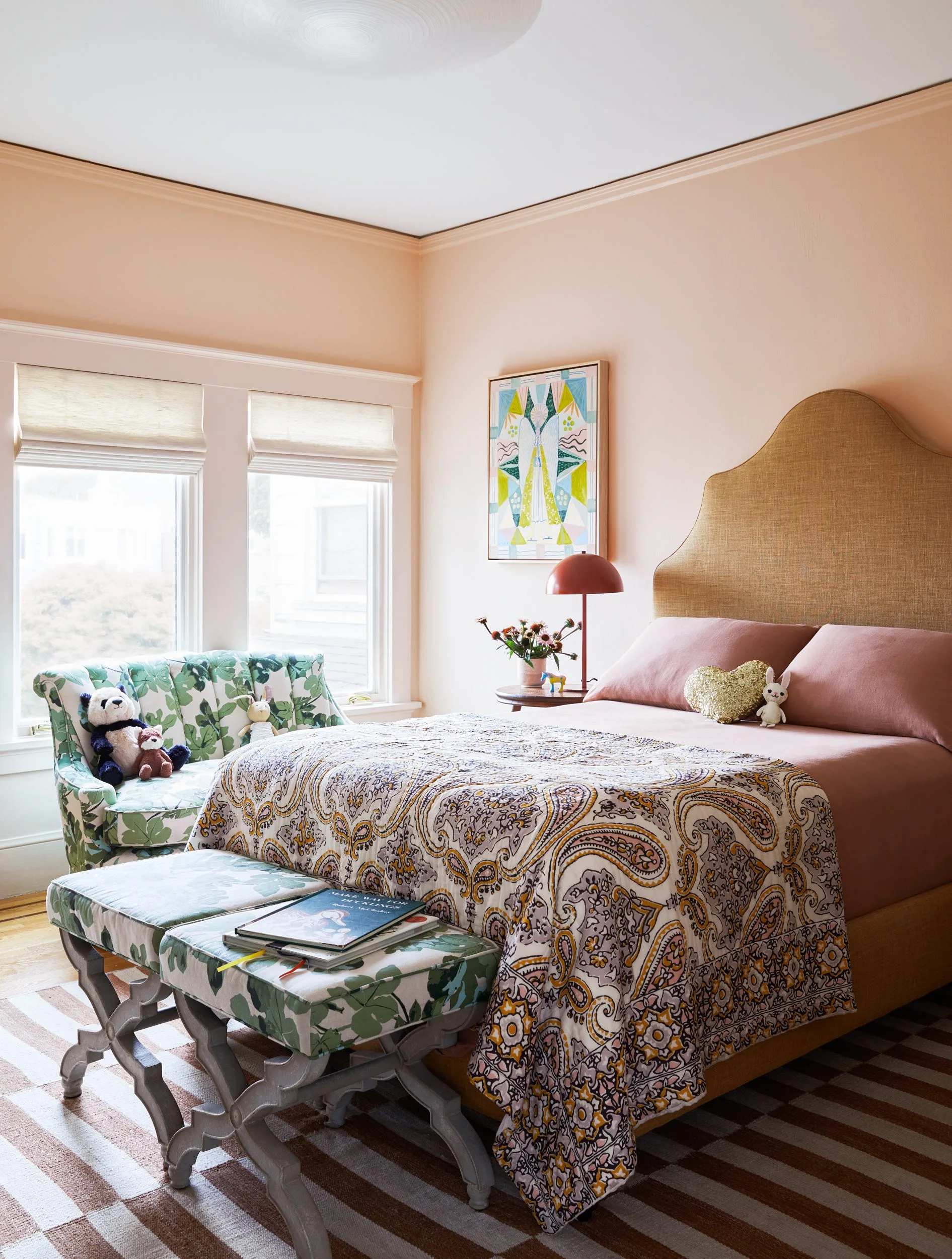 A kid's bedroom features peach walls, a high upholstered headboard, a green floral chaise, matching benches, and a brown striped rug.