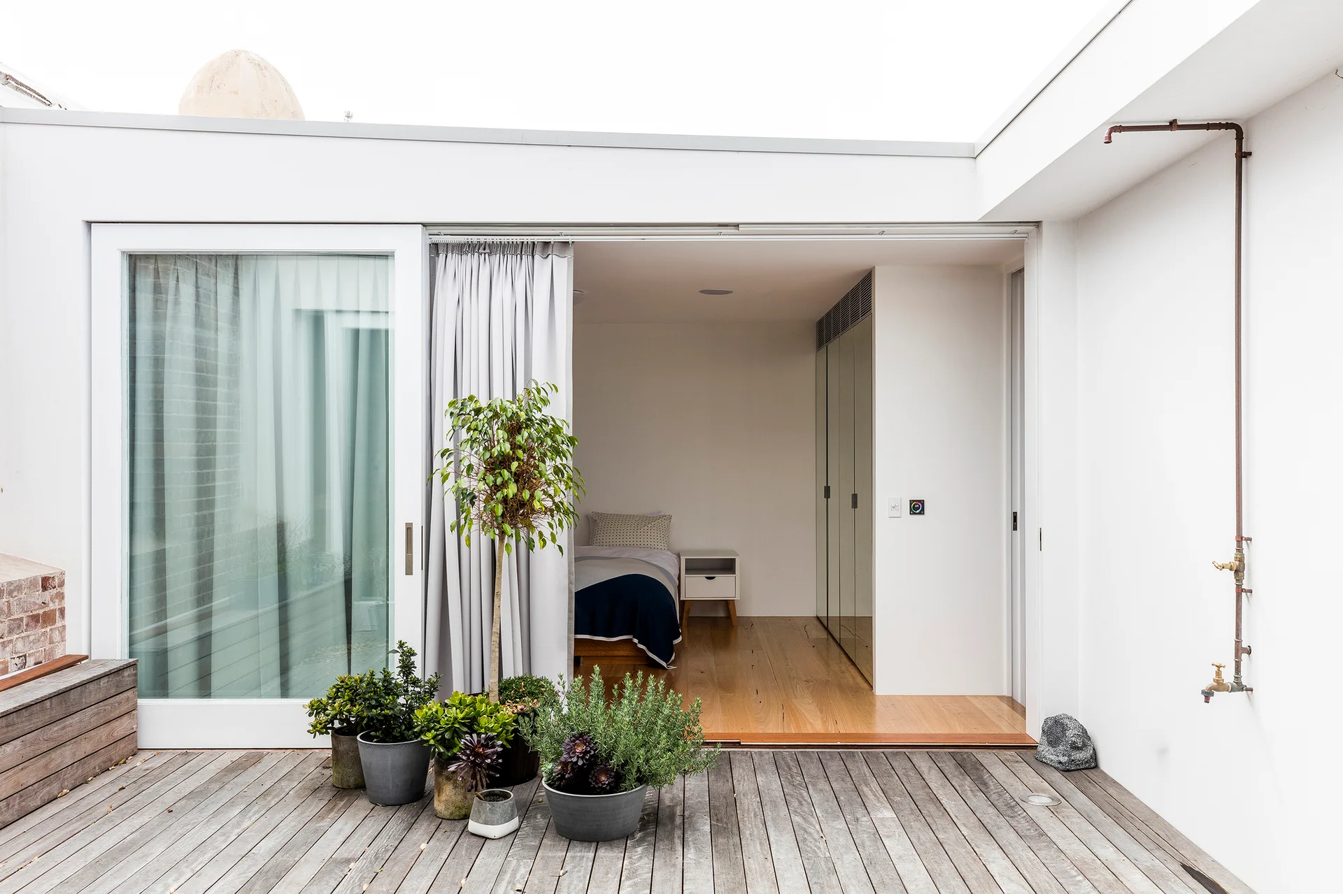 A weathered wooden patio with green potted plants leads through a sliding glass door into a minimalist bedroom with built-in closets.