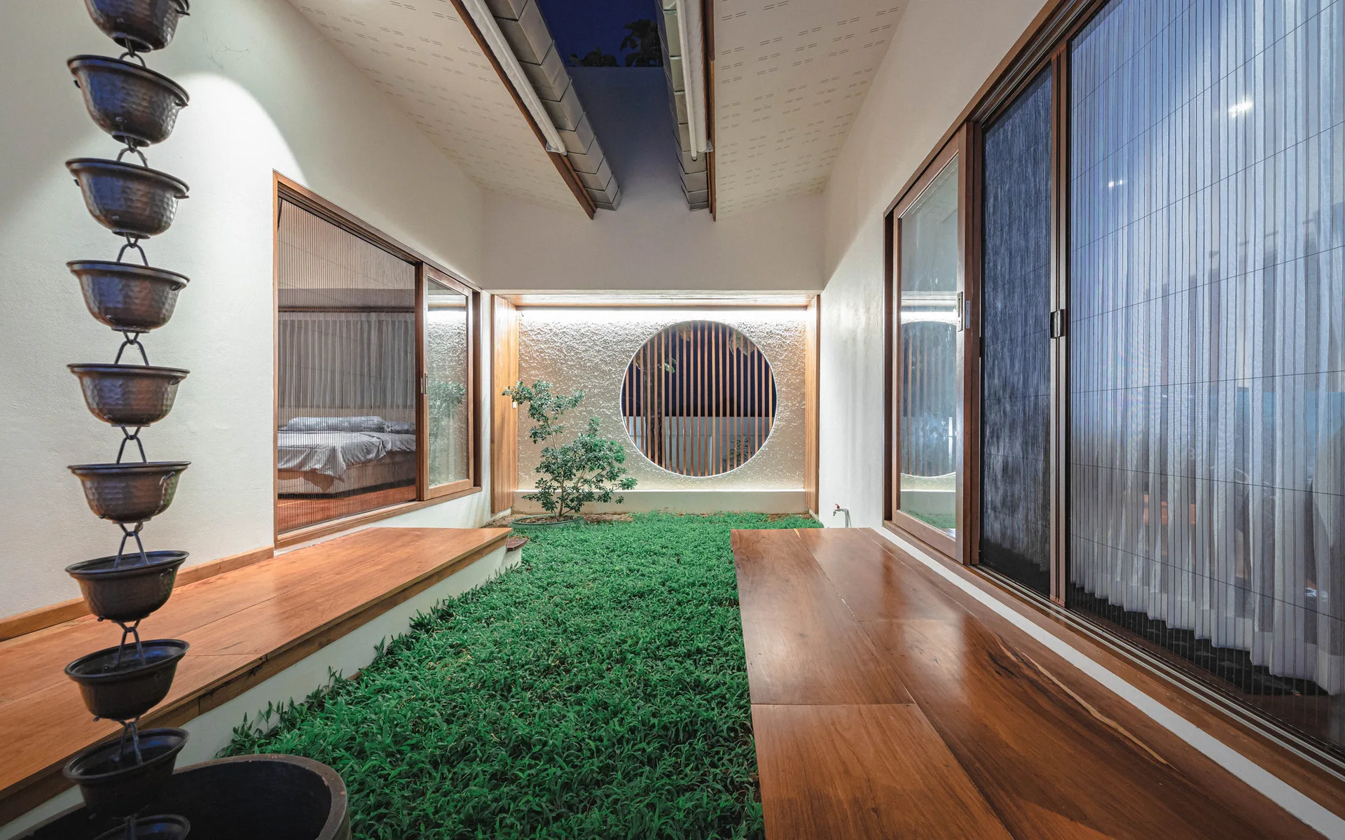 An enclosed courtyard features a central patch of green grass bordered by wooden decking, a textured back wall with a circular window, and a rain chain.