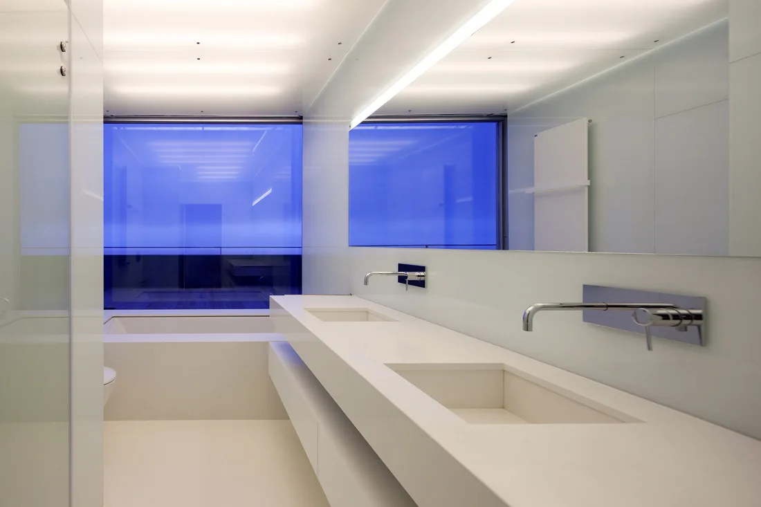 A minimalist white bathroom featuring a long solid-surface double vanity with integrated sinks, wall-mounted chrome faucets, and a large mirror.