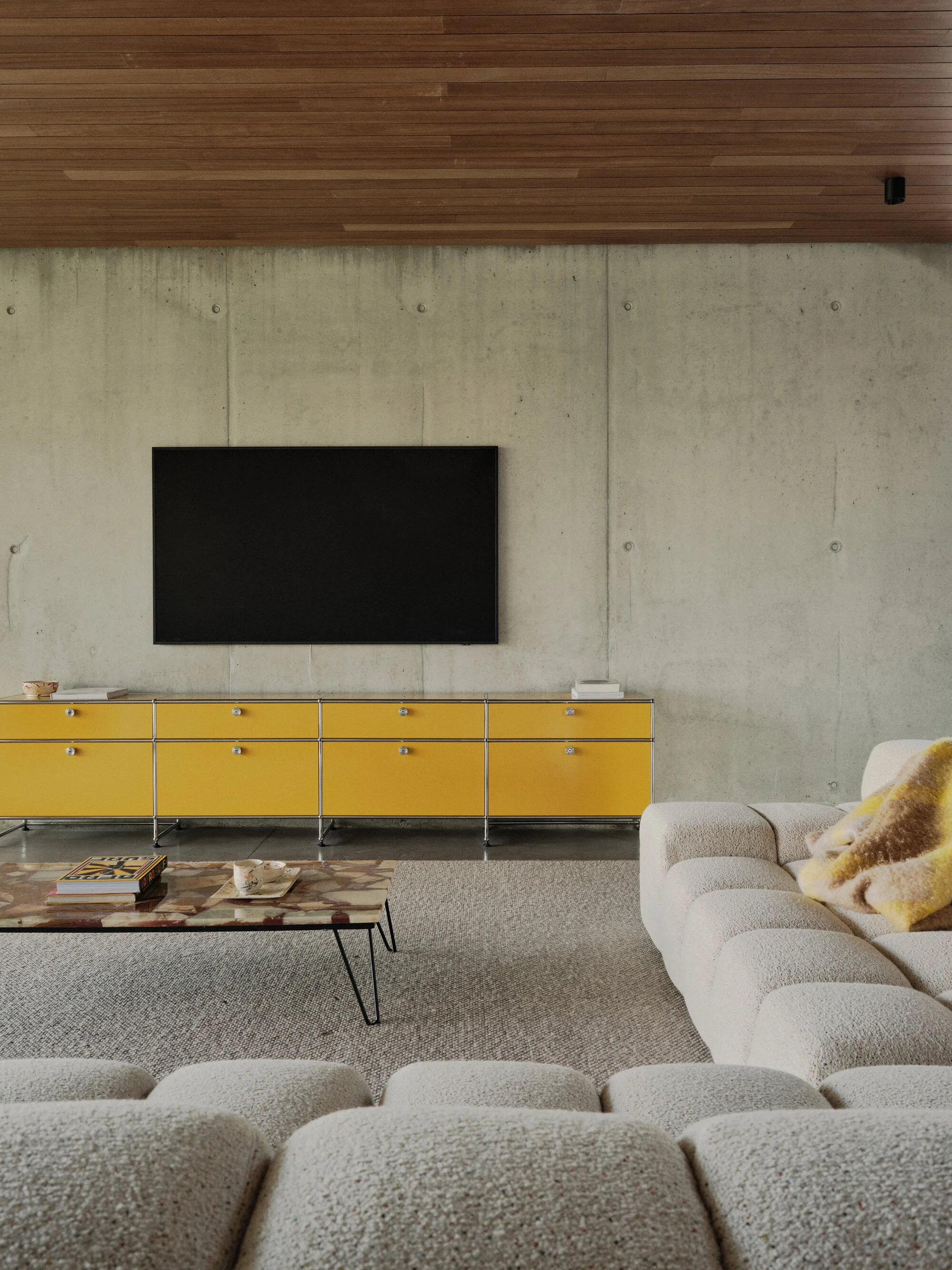 A modern living room features a cream sectional sofa, a bright yellow modular media console, a wall-mounted television, and a concrete wall.
