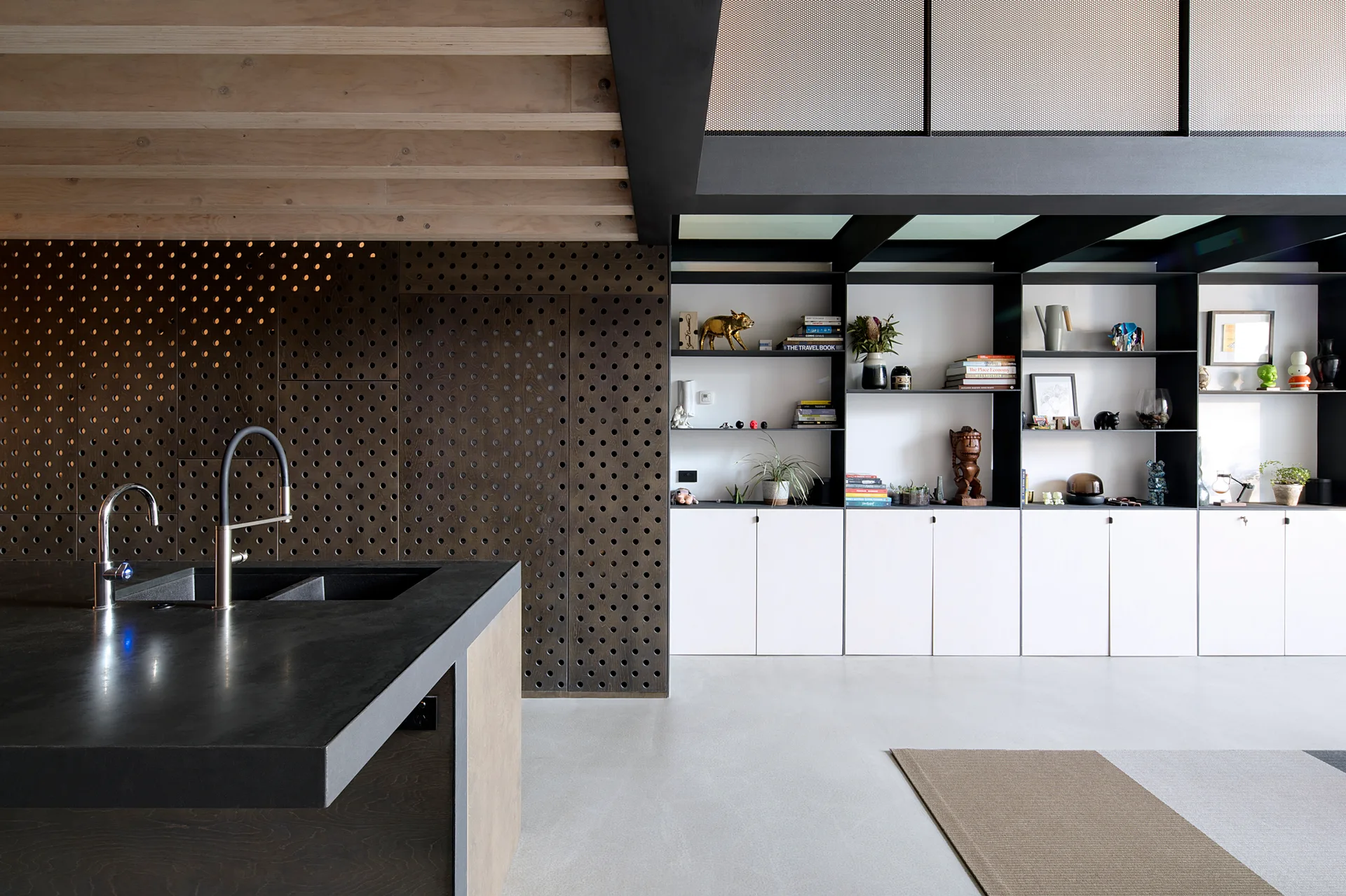 A modern kitchen features a dark island with a faucet, a perforated brown wood accent wall, and white built-in cabinets with open shelving.