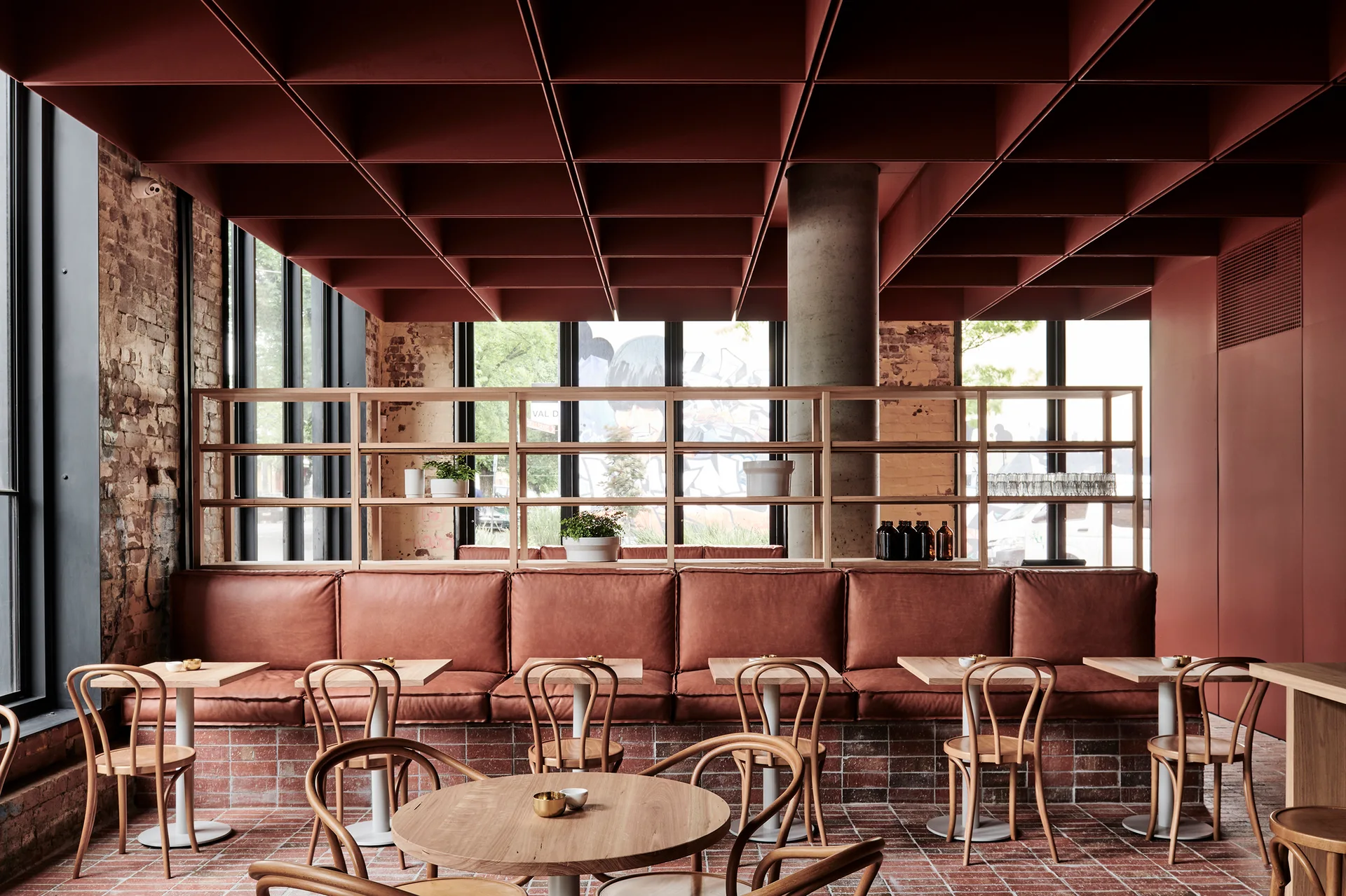 A modern cafe interior features a prominent red grid ceiling, leather banquette seating, wooden tables, and an open shelving divider.