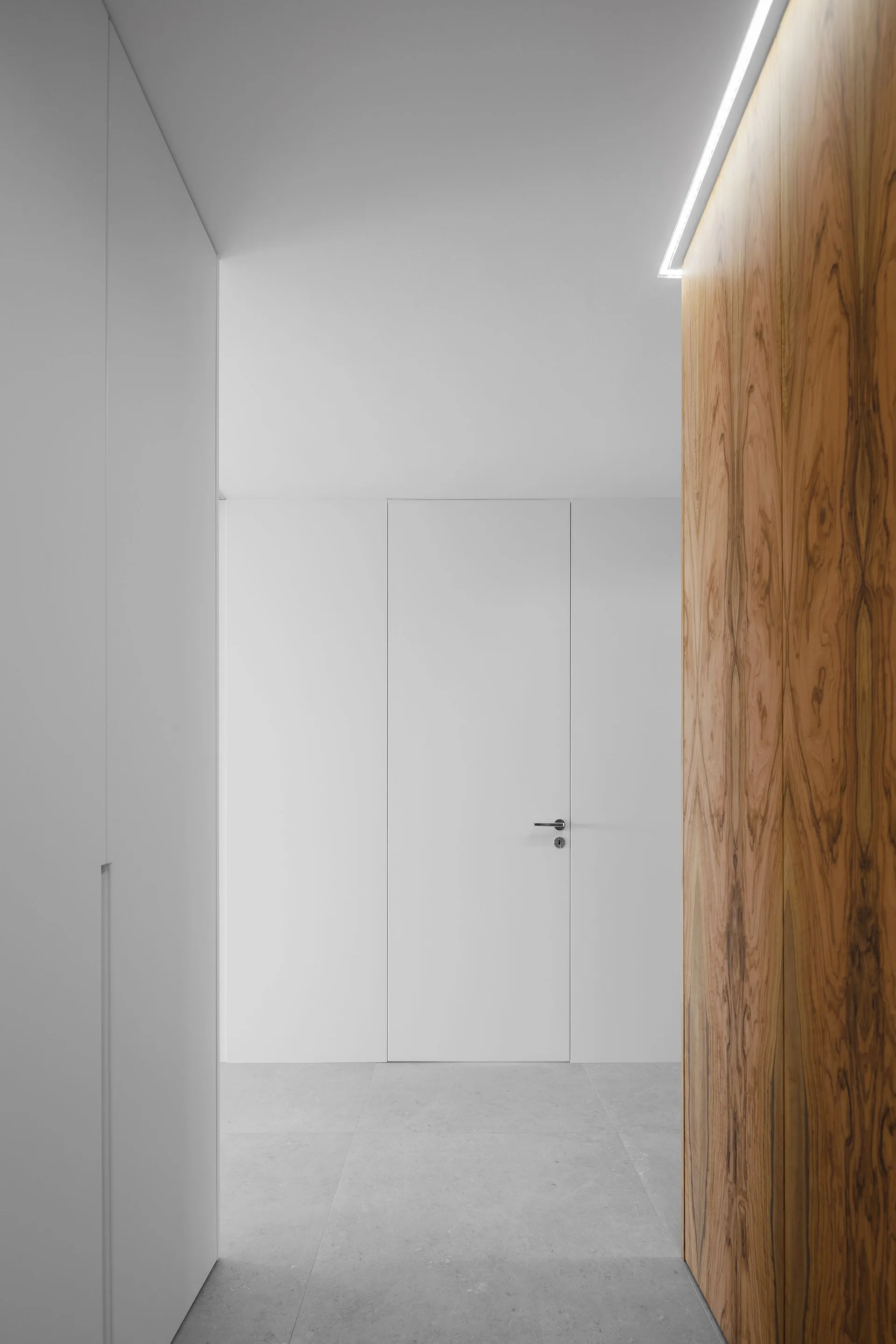 A minimalist hallway featuring a flush white door, large grey floor tiles, and a contrasting warm wood-paneled wall highlighted by overhead cove lighting.