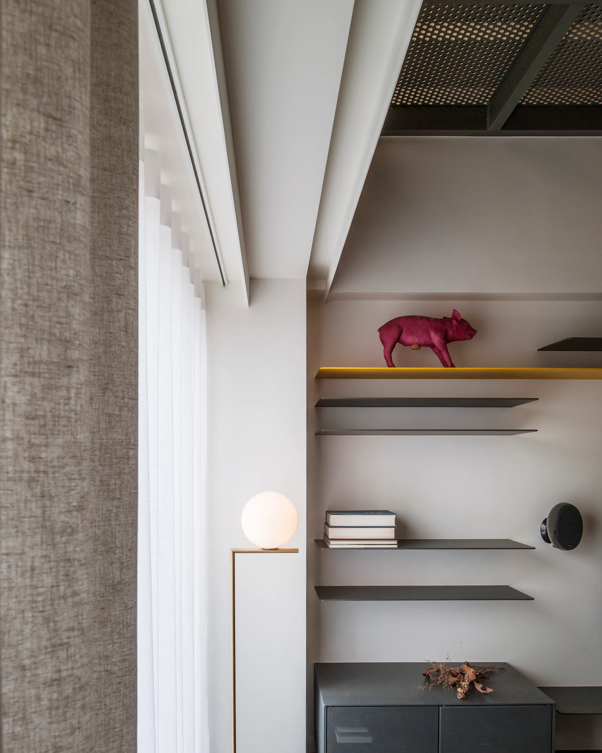 Minimalist grey wall featuring thin metal floating shelves with a magenta sculpture next to a spherical brass globe floor lamp and light drapes.