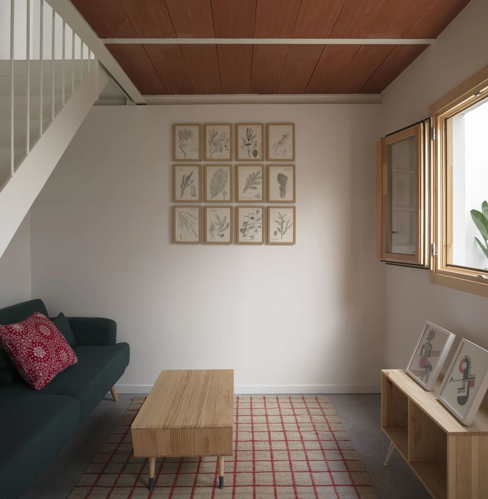 A minimalist living room features a dark sofa under a white staircase, a botanical grid gallery wall, and a wood plank ceiling.