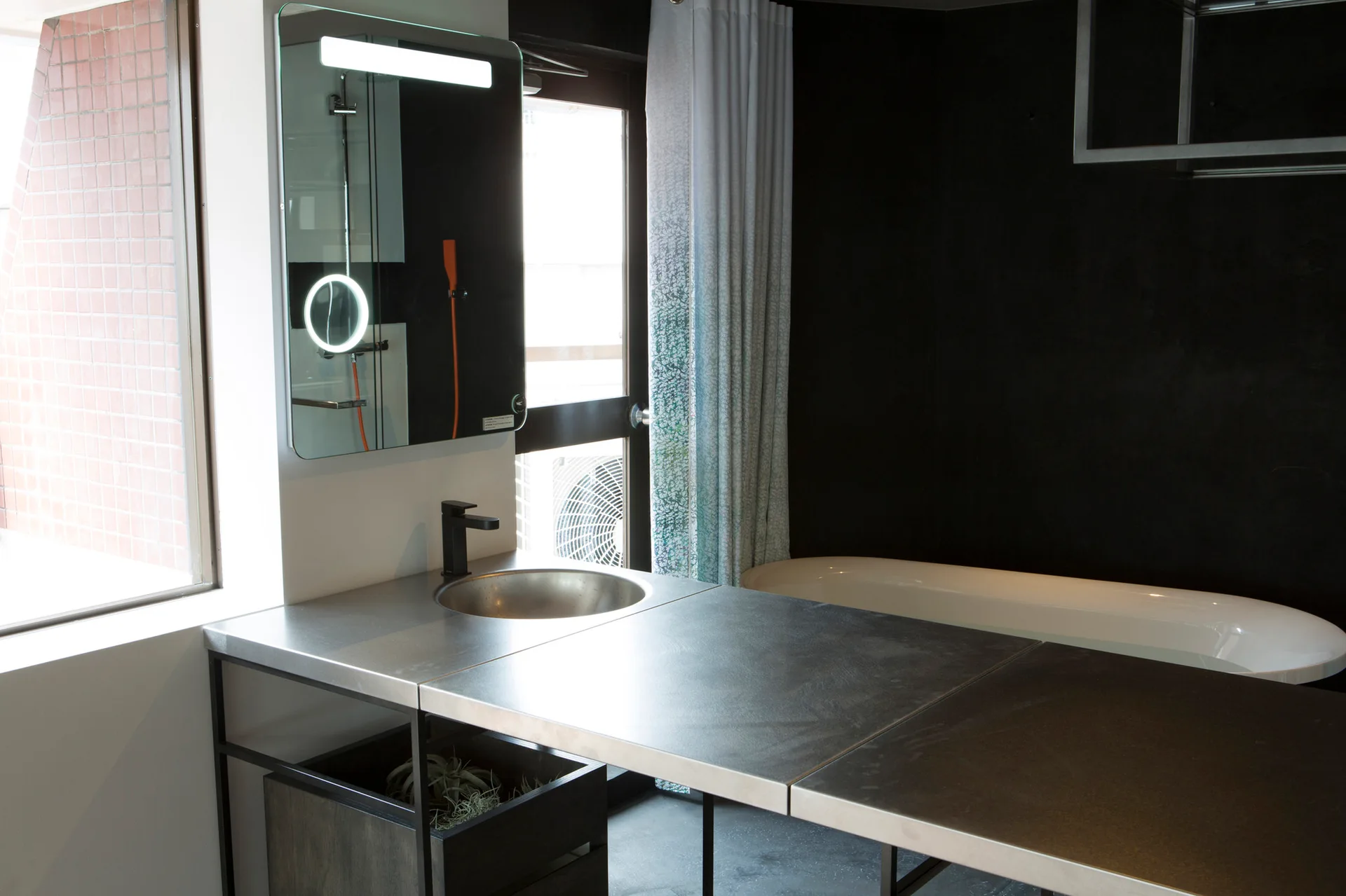 An industrial bathroom with a metal countertop vanity, a matte black faucet, an illuminated mirror, and a white tub against a black wall.