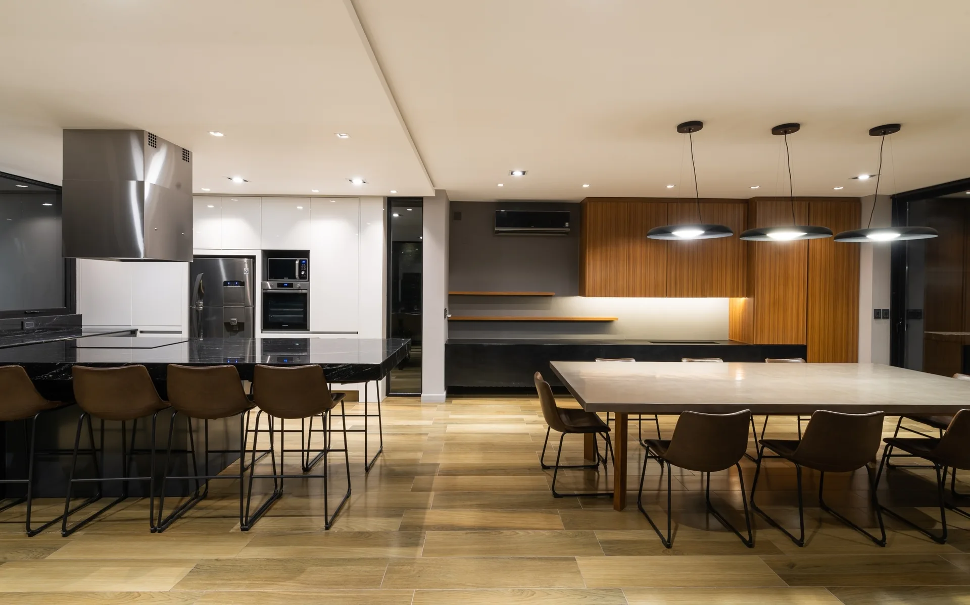 A modern open-plan kitchen and dining area featuring a black stone island, wood-toned cabinetry, a long dining table, and modern pendant lights.