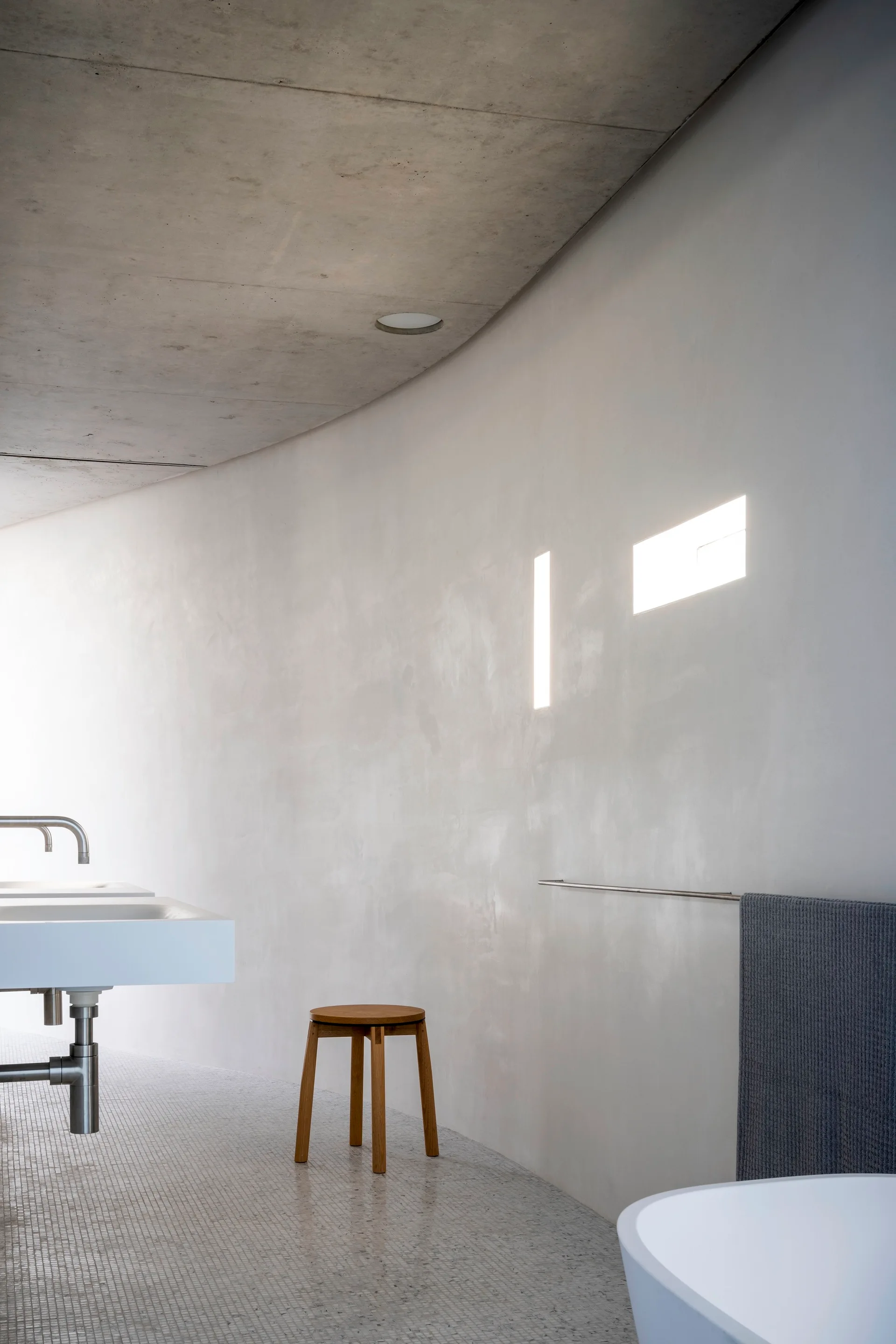 A minimalist bathroom featuring a curved plaster wall, an exposed concrete ceiling, a white wall-mounted sink, and a wooden stool.