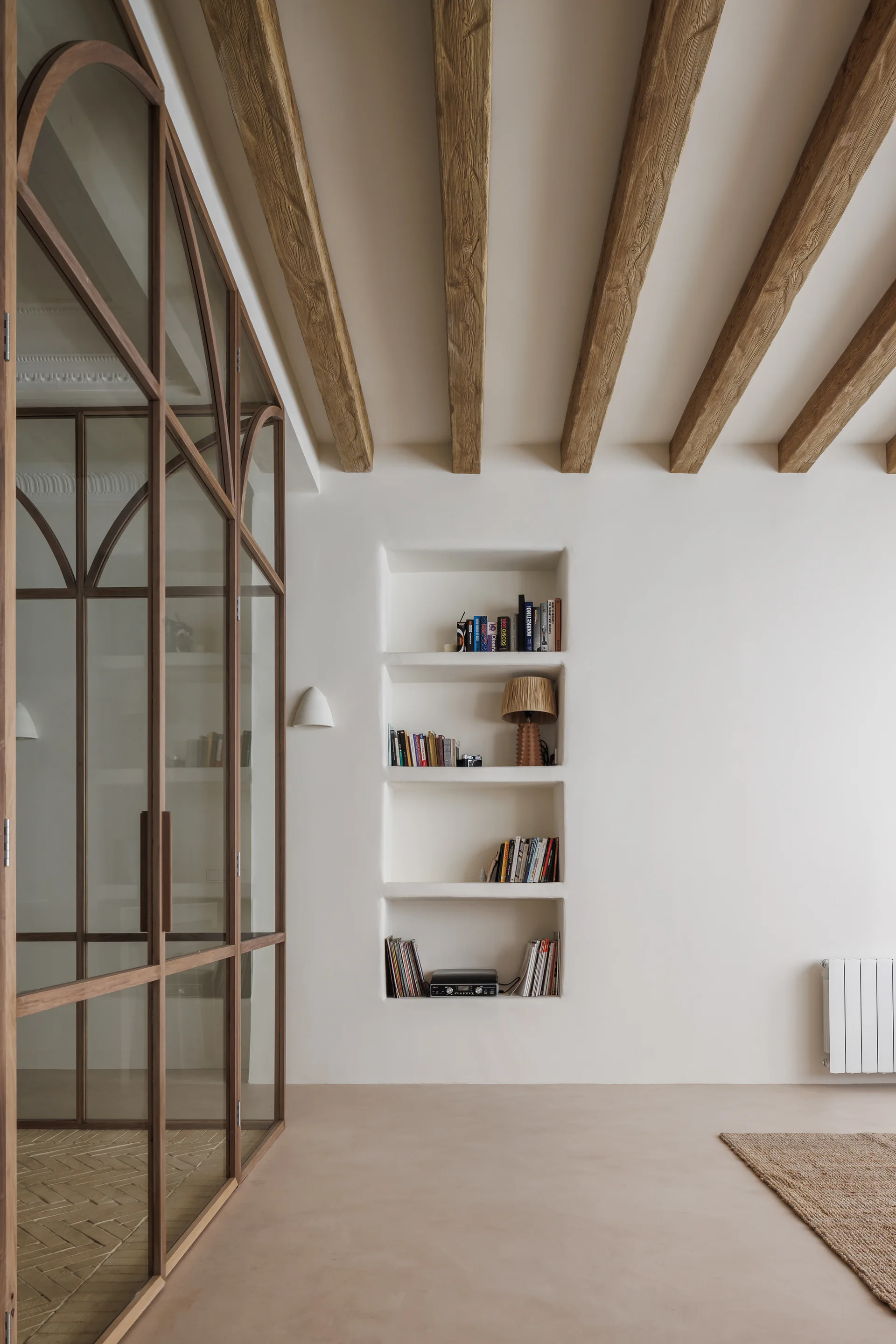 A minimalist living space features a large wood-framed arched glass partition, recessed wall shelves filled with books, and exposed wooden ceiling beams.