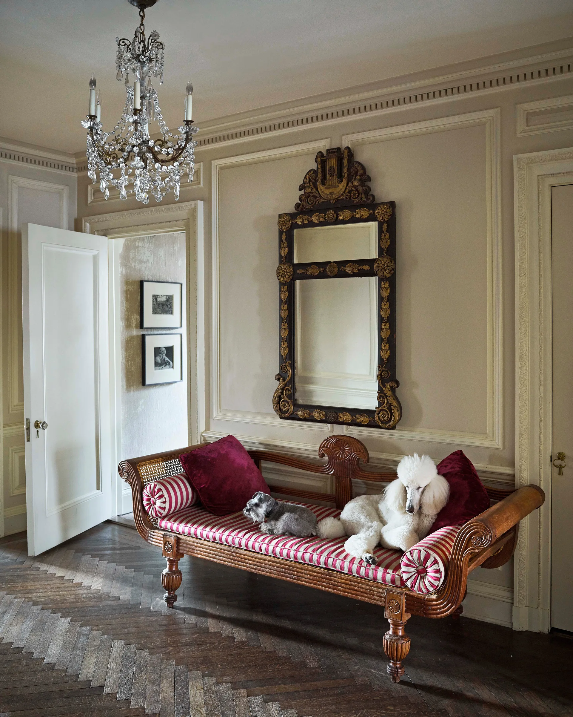 An entryway features a striped wooden settee with red velvet pillows beneath an ornate mirror, set on dark herringbone parquet flooring.