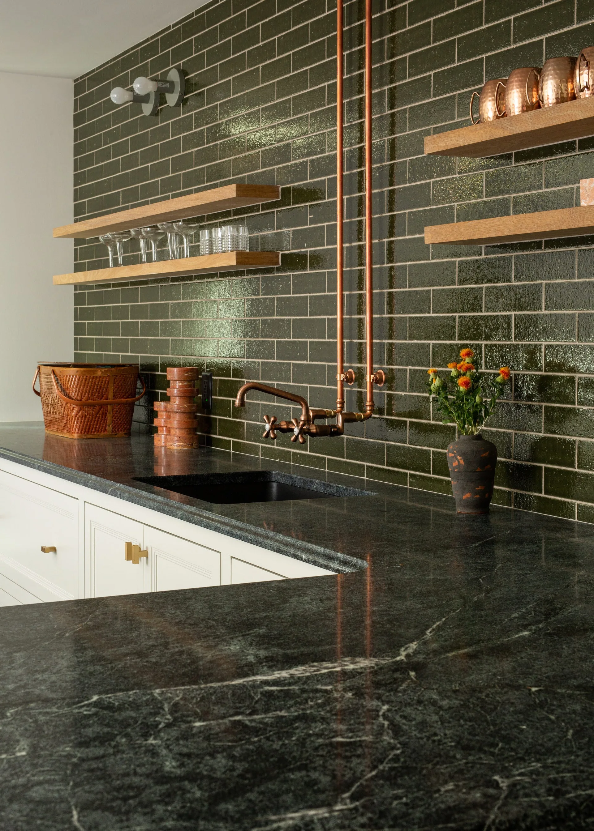 A wet bar features a glossy green subway tile backsplash, exposed copper piping and faucet, dark veined stone countertops, and floating wood shelves.