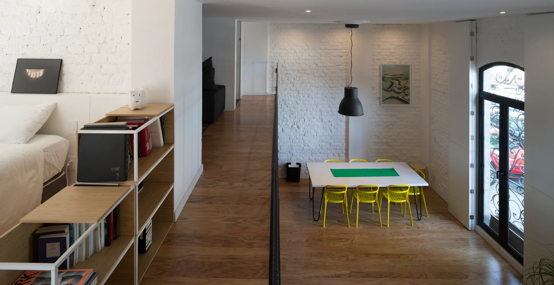 A split-level loft interior features a white dining table with yellow chairs beneath a black pendant light, adjacent to a raised bedroom area.