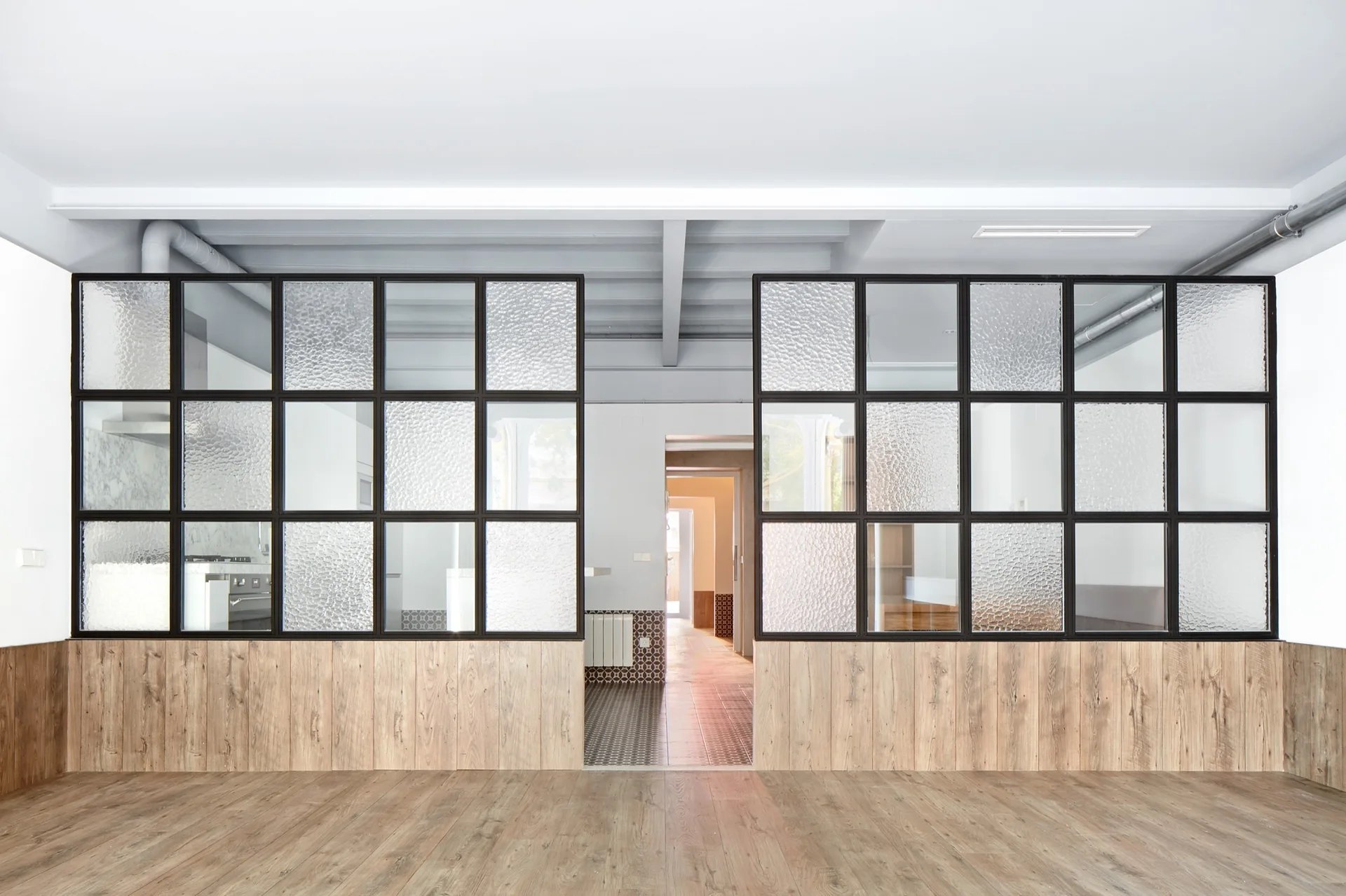 A black-framed glass partition wall with textured panes sits above wood wainscoting next to an open hallway.