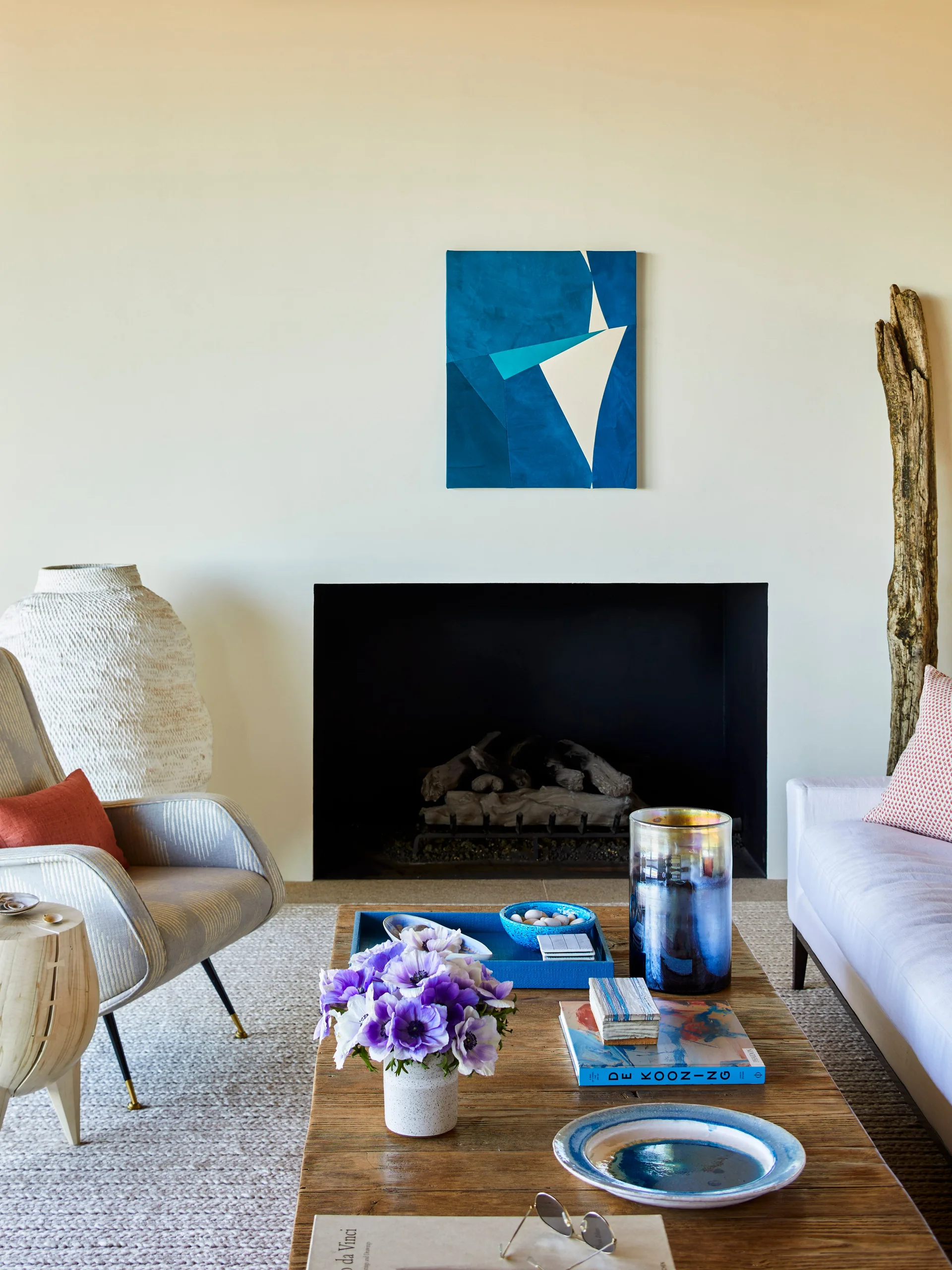 A living room features a black inset fireplace, a blue abstract painting, a striped armchair, and a wooden coffee table with books and purple flowers.