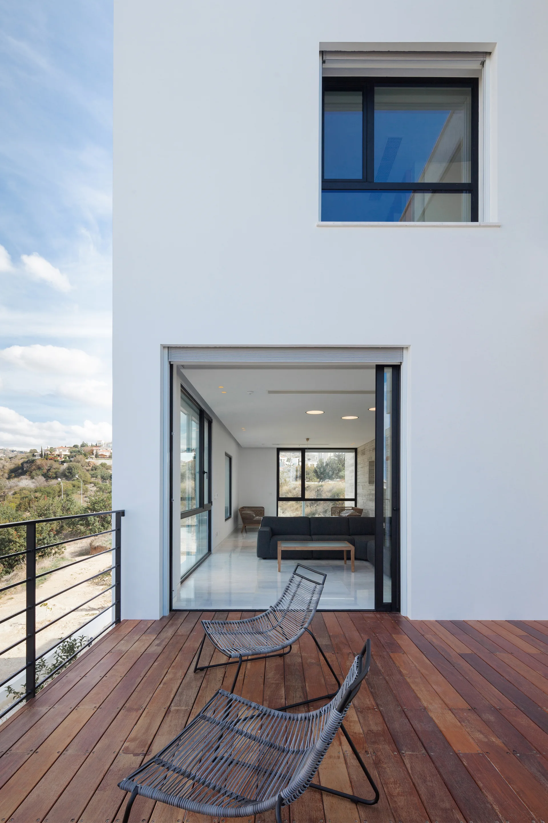 Two gray wire-frame lounge chairs sit on a rich wood plank deck next to an open sliding glass door showing a modern living room.