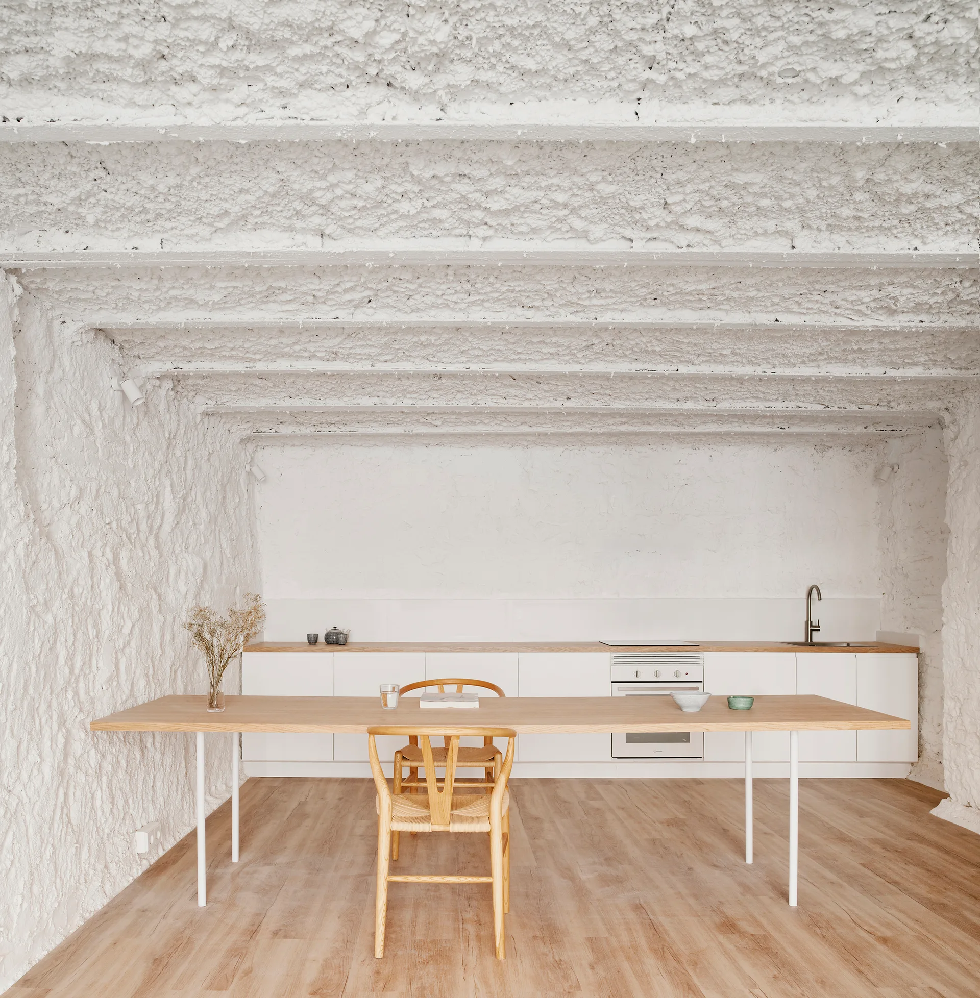 A minimalist kitchen features a long light wood dining table with a wishbone chair set against heavily textured white plaster walls and ceiling.