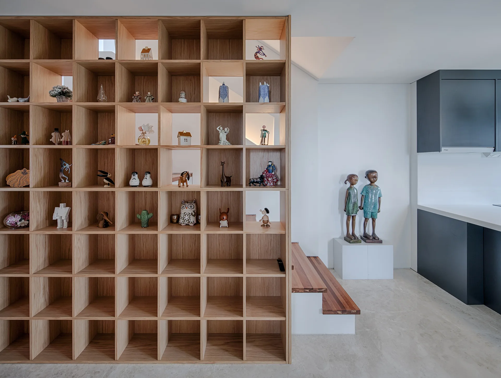 A floor-to-ceiling light wood grid shelving unit partitions a room beside a staircase, displaying small figurines in its open square cubbies.