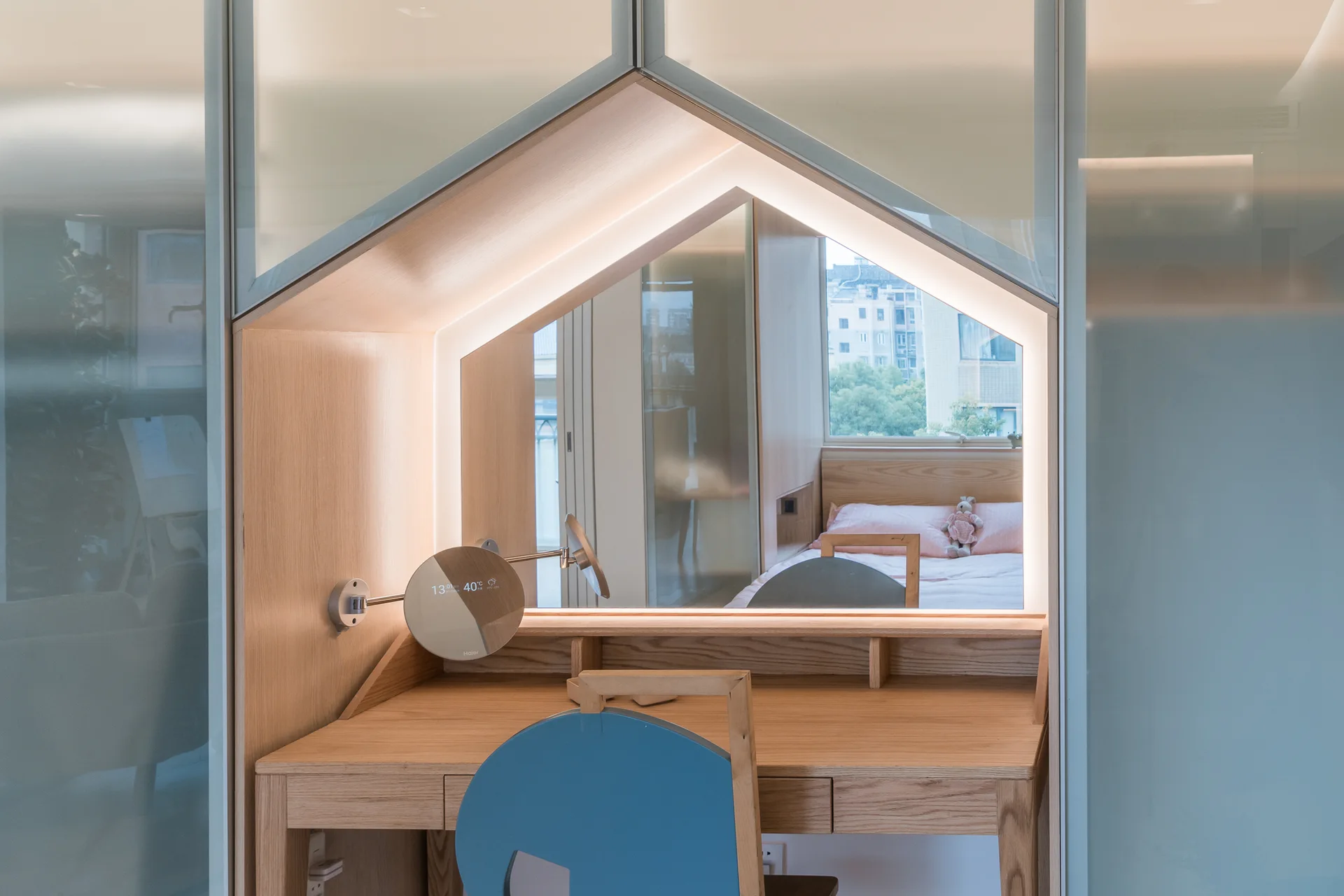 A light wood built-in vanity desk featuring a house-shaped mirror outlined with LED strip lights and a blue wooden chair in a bedroom.