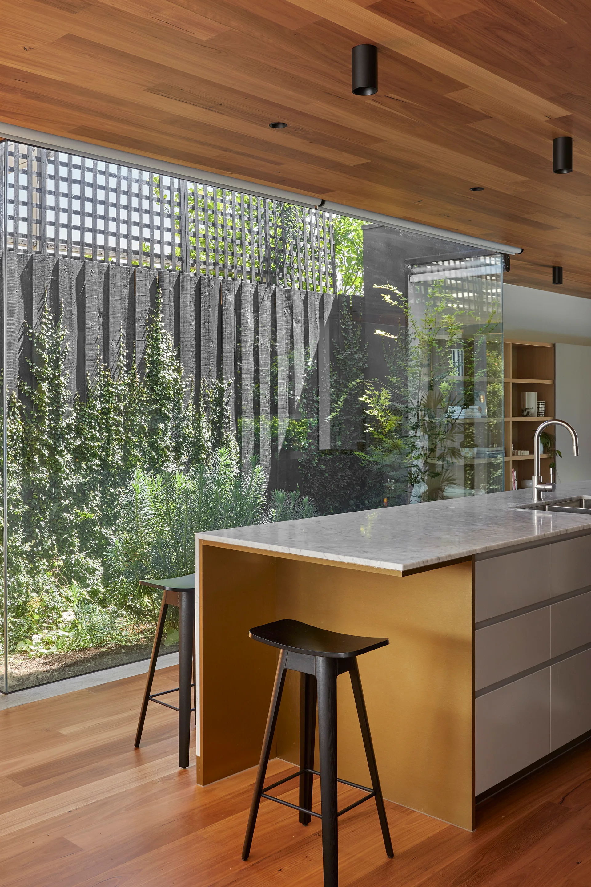 A modern kitchen island with a gold-toned side panel and marble countertop stands before a large window overlooking a garden.