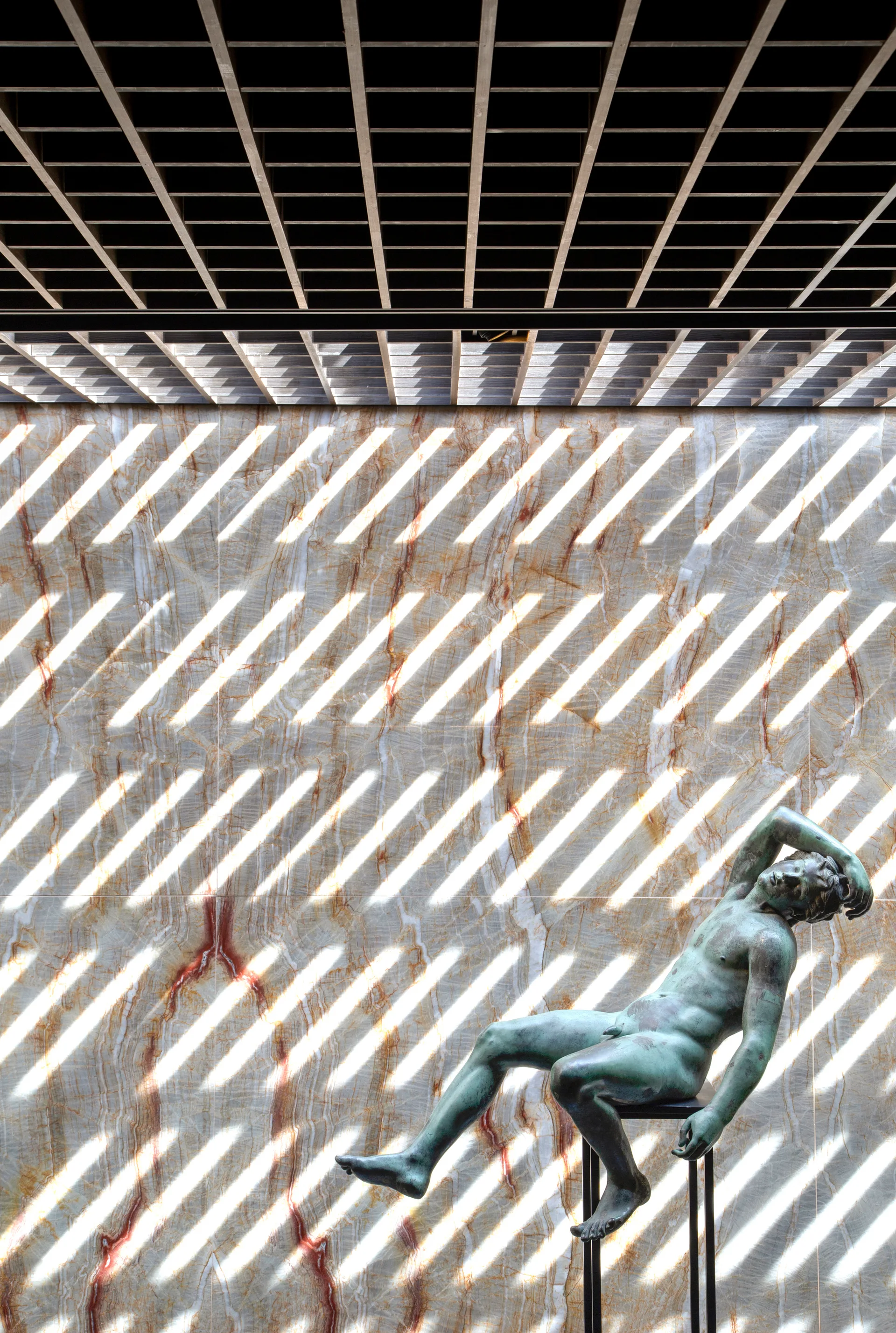 A green bronze statue sits on a thin pedestal against a tan veined stone wall illuminated by striped light patterns from a black grid ceiling.