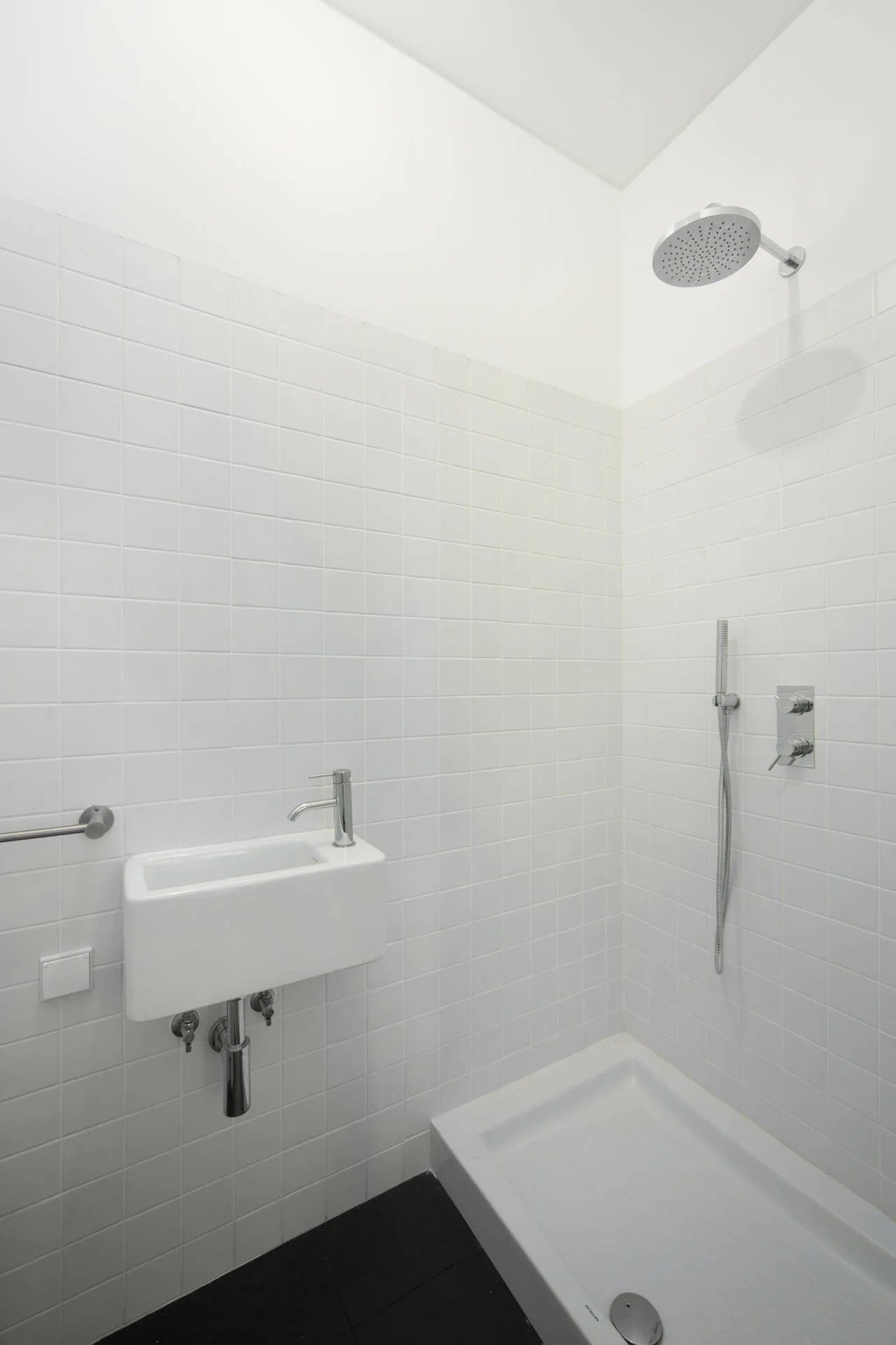 A minimalist bathroom featuring white square grid tile walls, a small floating sink, black floor tile, and a walk-in shower.