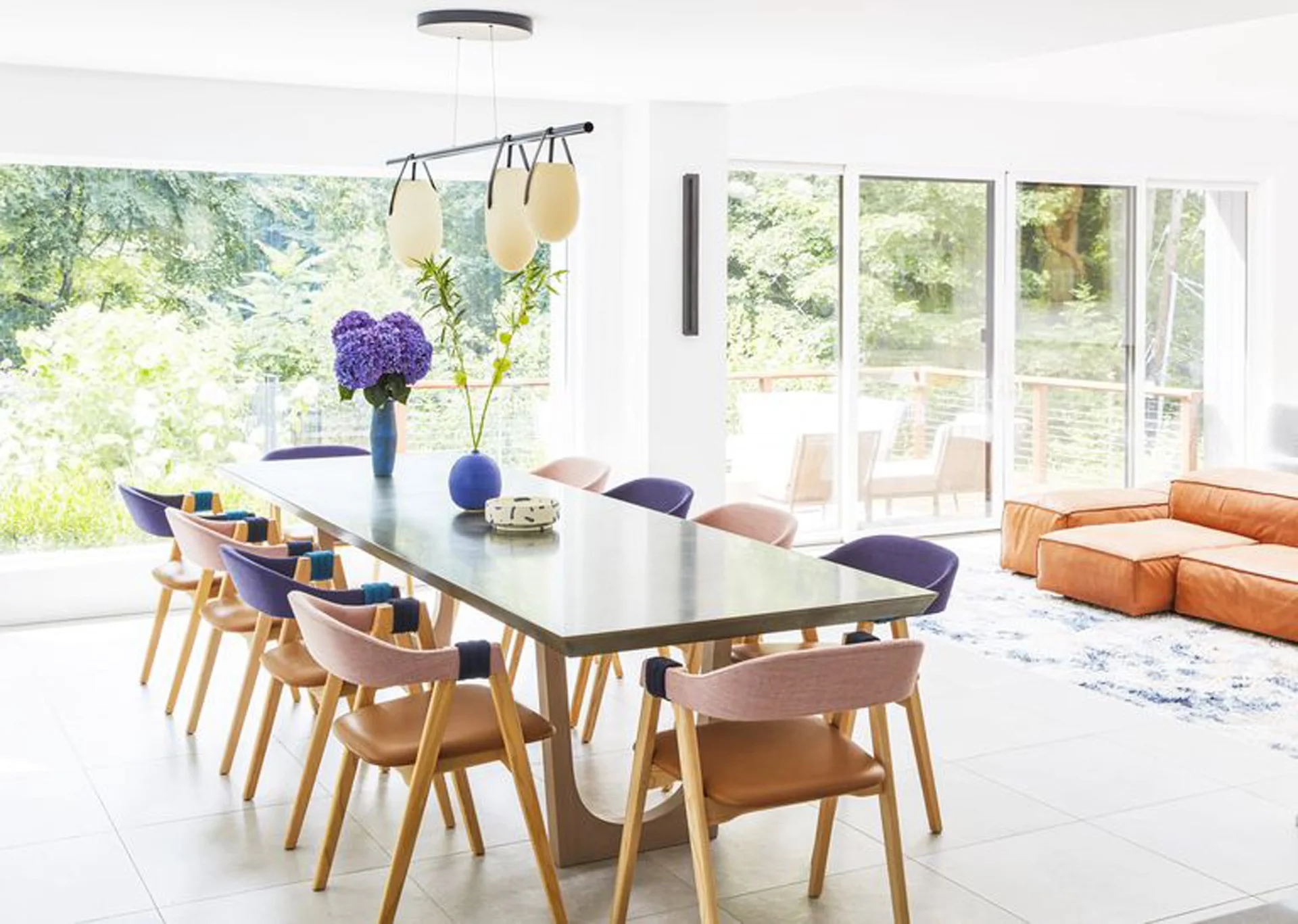 A long dining table surrounded by purple and pink upholstered chairs stands in a bright room with sliding glass doors and a leather sofa.