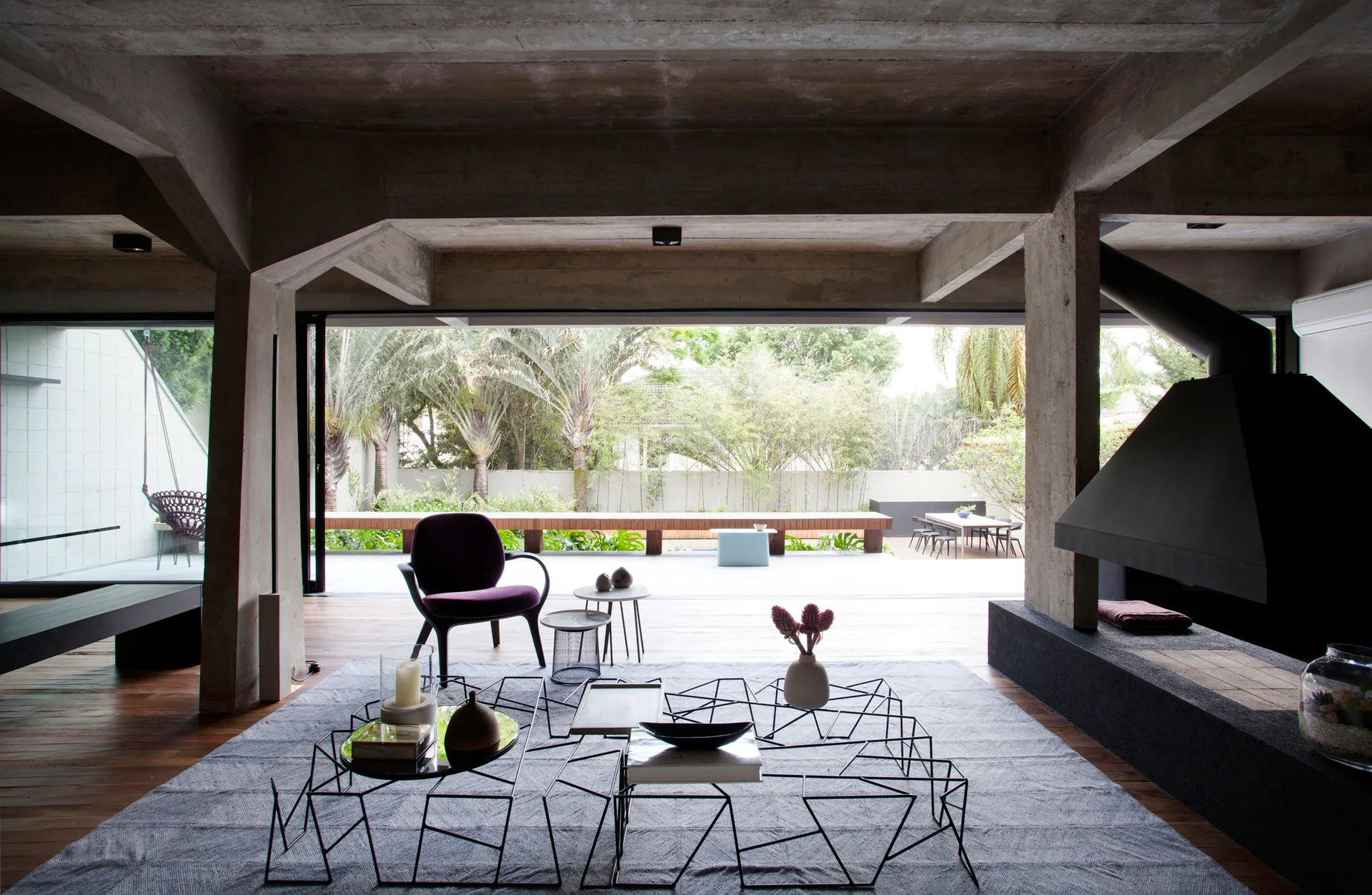 A modern living room features raw concrete ceilings, a black fireplace, a purple armchair, and sliding glass doors opening to a lush green patio.