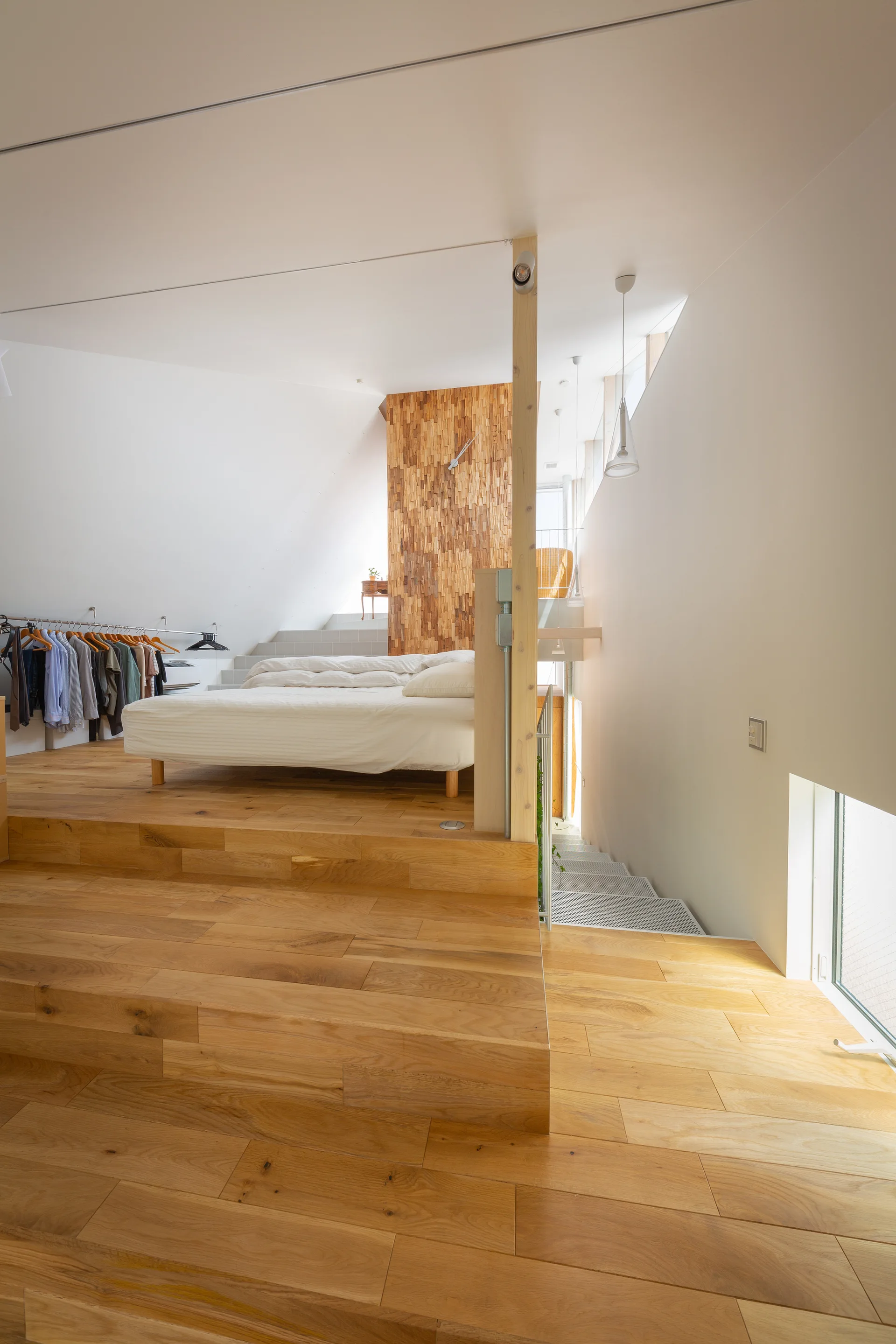 A minimalist bedroom on an elevated wood platform features a textured wood-block accent wall, a white bed, and an adjacent metal staircase.