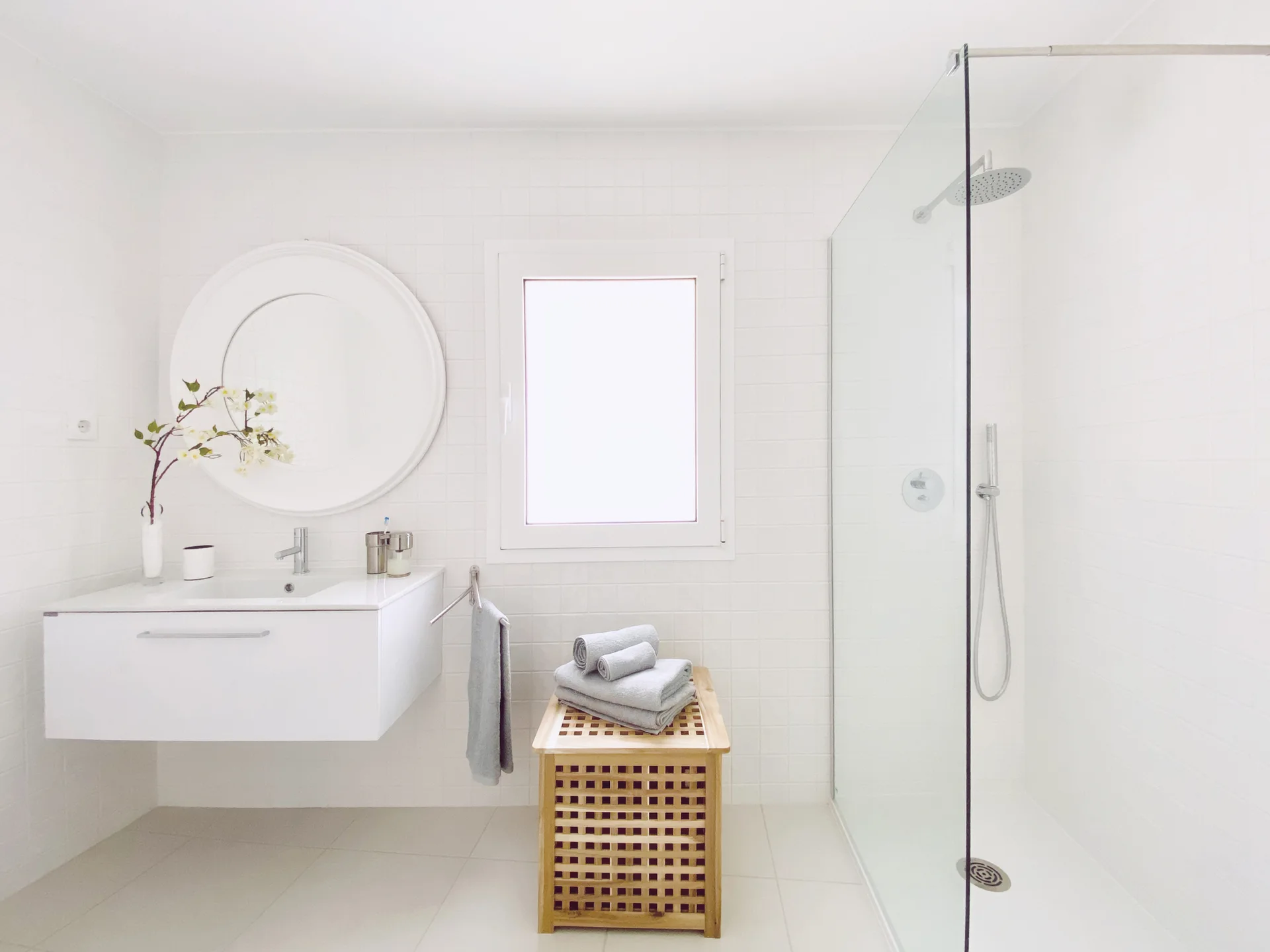 A minimalist white bathroom featuring a floating vanity under a round mirror, a glass-enclosed walk-in shower, and a slatted wooden bench.